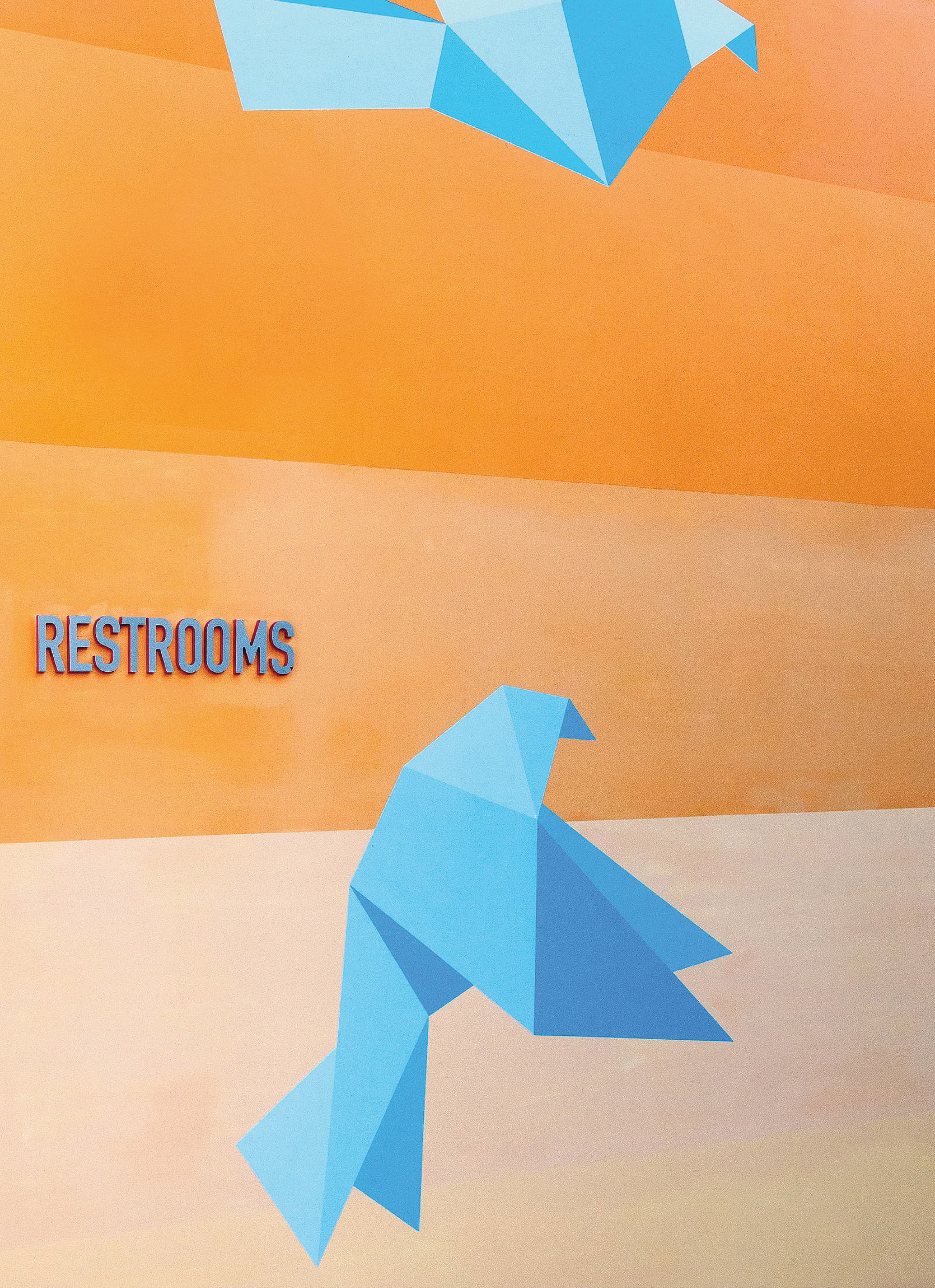 Restroom wayfinding signage integrated into a wall mural featuring geometric origami birds in blue and orange, blending environmental graphic design and experiential graphics for a hospitality or mixed-use environment by RSM Design.