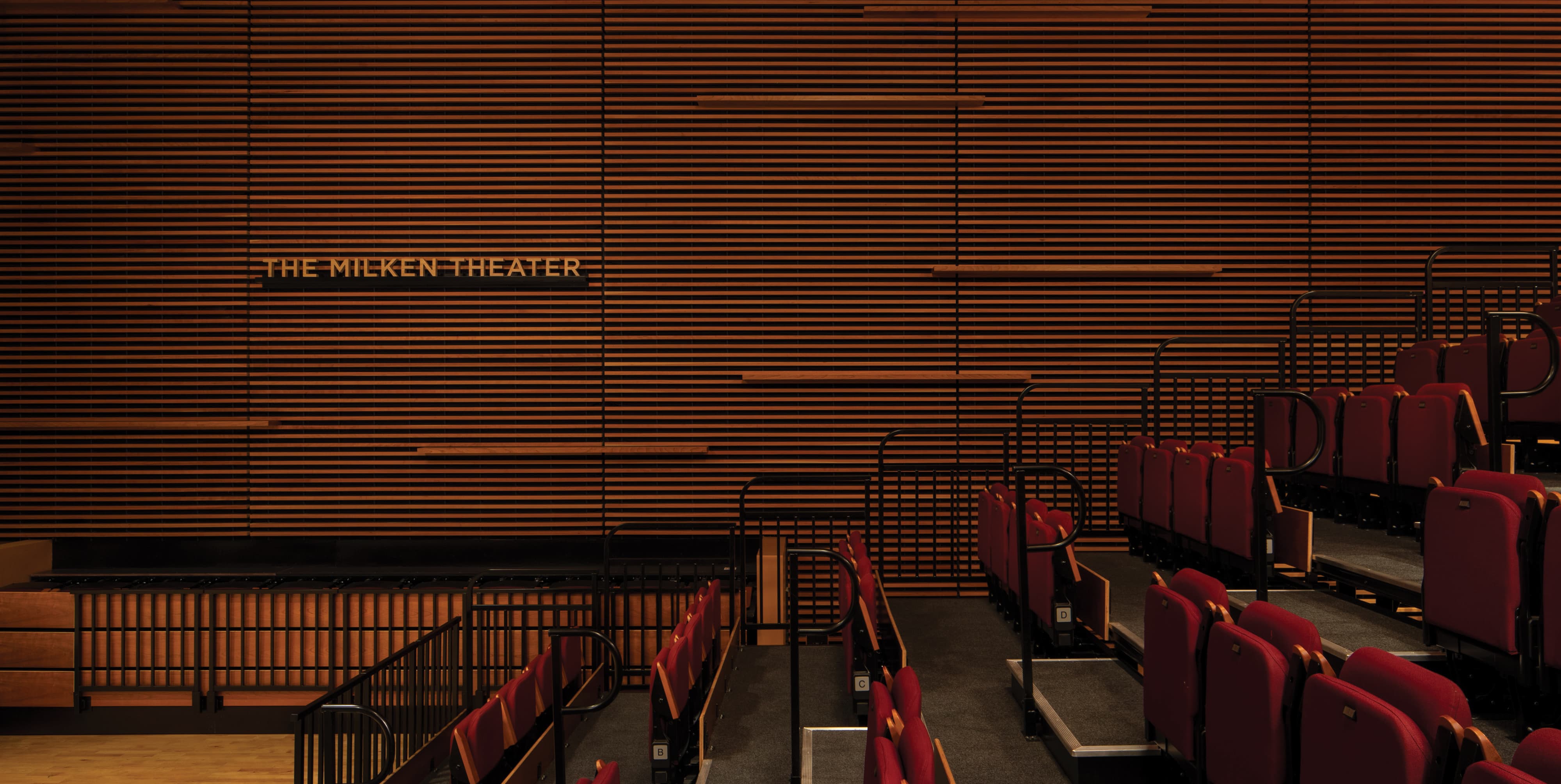 Interior of The Milken Theater featuring warm gold dimensional donor recognition lettering mounted flush within a floor-to-ceiling horizontal wood slat acoustic wall, overlooking rows of deep red upholstered theater seats arranged in a tiered auditorium configuration — exemplifying RSM Design's donor recognition and specialty graphics services that integrate naming opportunities with refinement and architectural sensitivity within the education and cultural markets