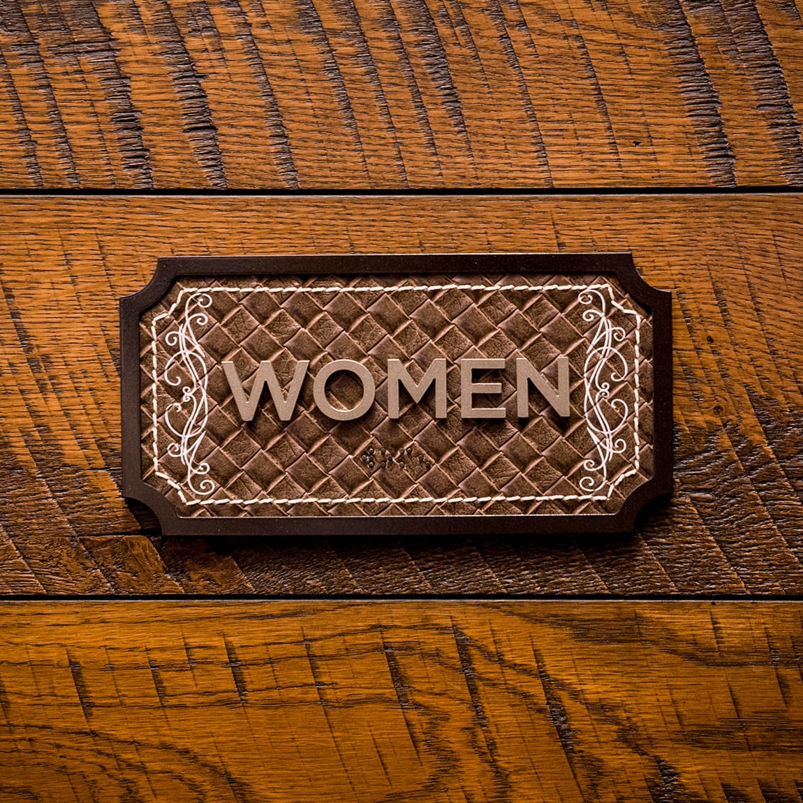 Hotel Drover women's restroom sign featuring tooled leather texture, decorative scroll detailing, and raised lettering mounted on a rustic wood wall, reflecting the hotel's Western-inspired interior design