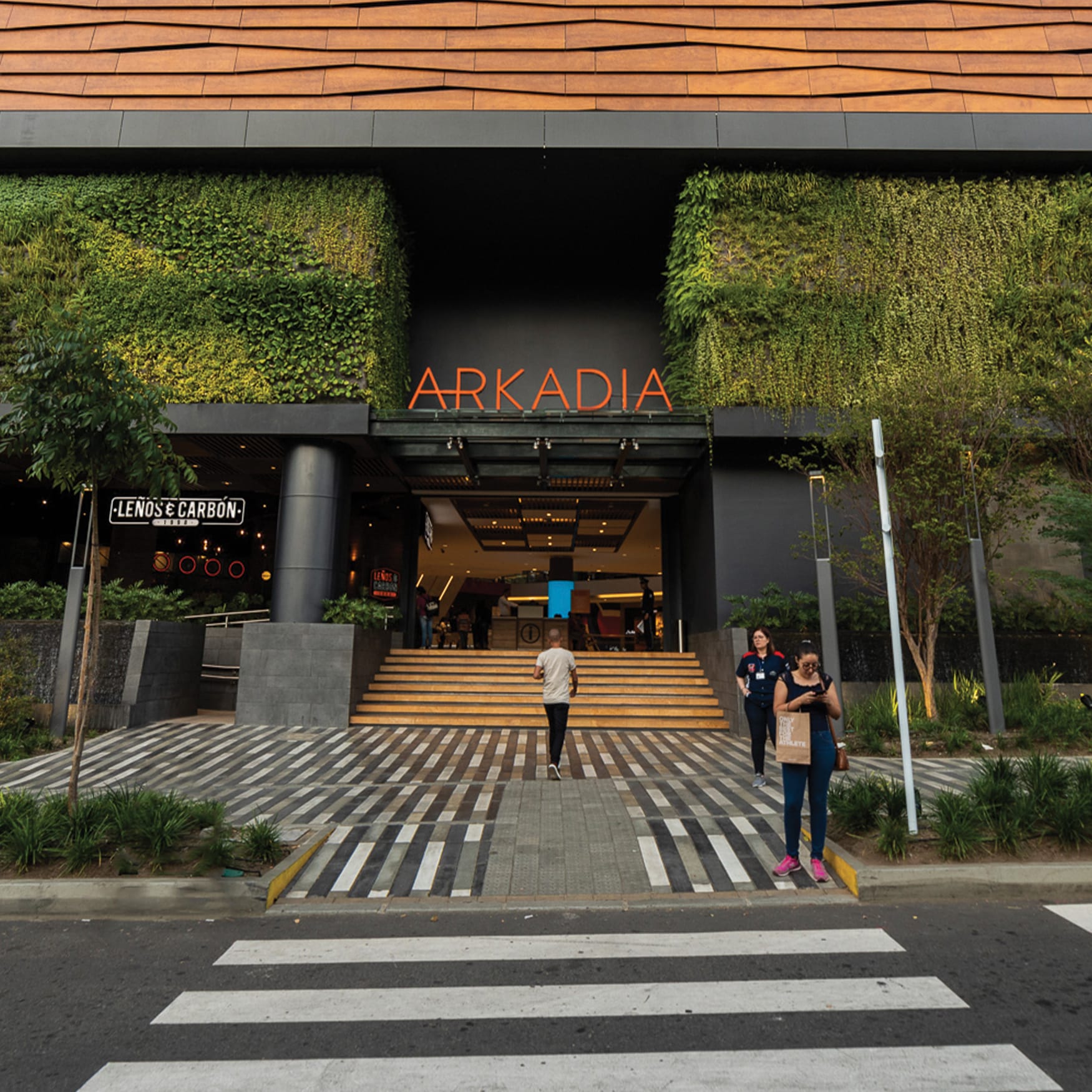 Modern commercial building entrance featuring the illuminated “ARKADIA” sign, lush green walls, contemporary landscaping, patterned pedestrian paving, and a restaurant storefront in a tropical urban setting.