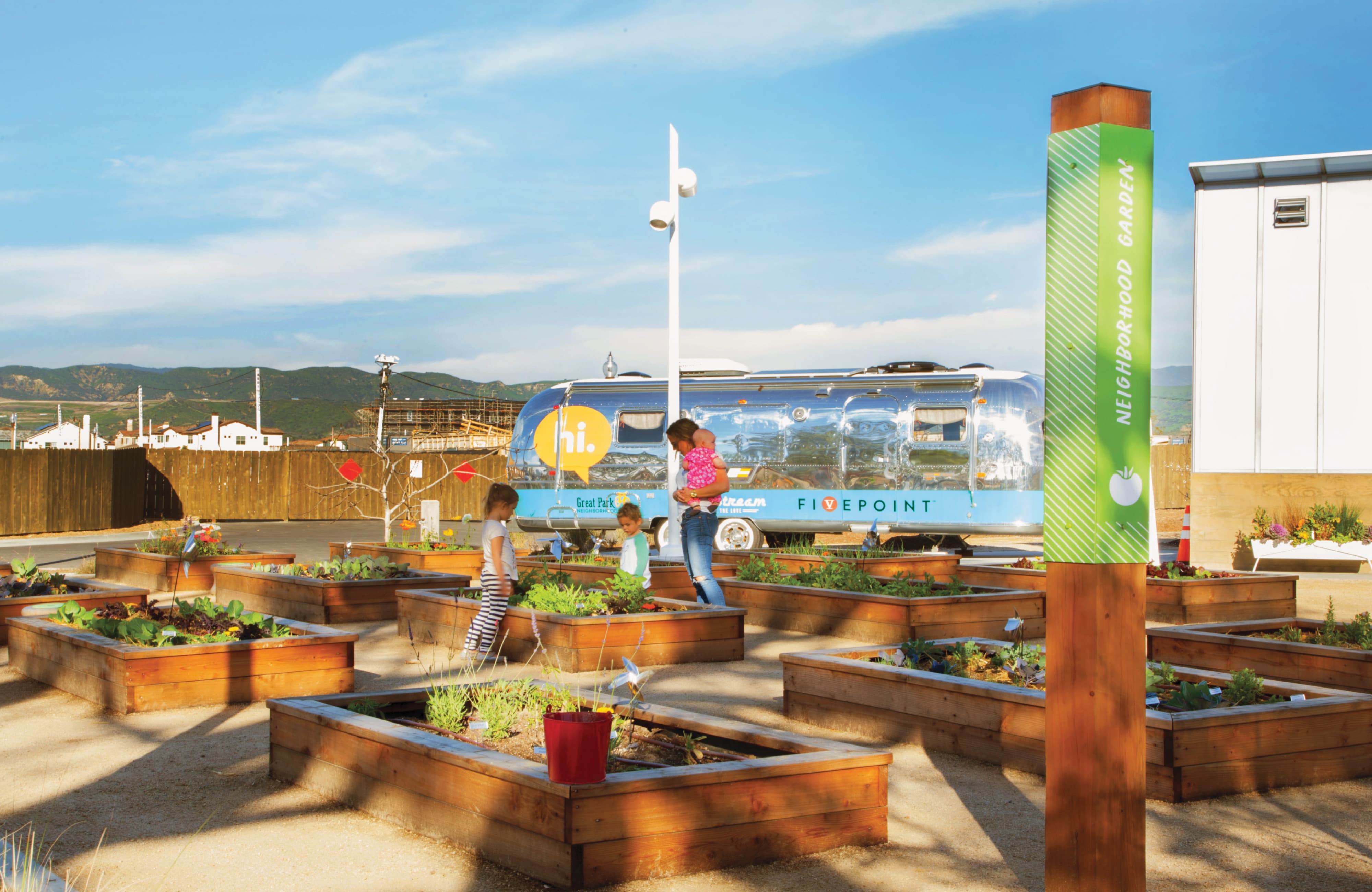 RSM Design wayfinding signage and placemaking for FivePoint at Great Park Neighborhoods in Irvine, California, featuring a branded green Neighborhood Garden identification post sign alongside raised wood planting beds with children and a parent exploring the community garden, with a FivePoint-branded Airstream trailer serving as an experiential branded environment activation in the background against a mountain landscape, demonstrating how environmental graphic design, specialty graphics, visual identity, and placemaking create meaningful, human-centered community connections for residential community, park, and mixed-use markets.