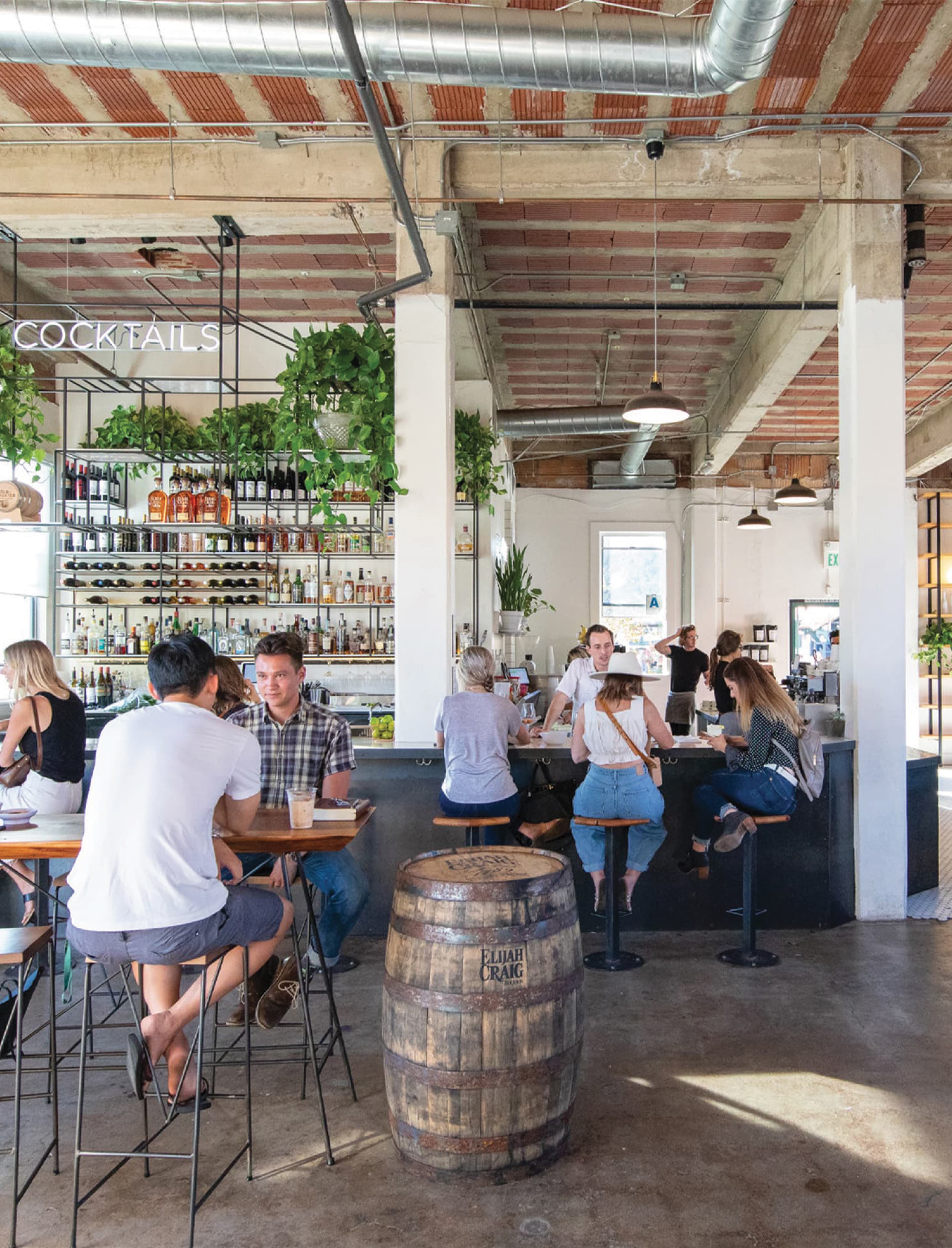 Industrial-chic bar interior with exposed brick ceiling, a neon 'Cocktails' sign, shelves of liquor bottles framed by hanging greenery, and patrons seated at high-top tables and the bar, with an Elijah Craig whiskey barrel repurposed as a table in the foreground.