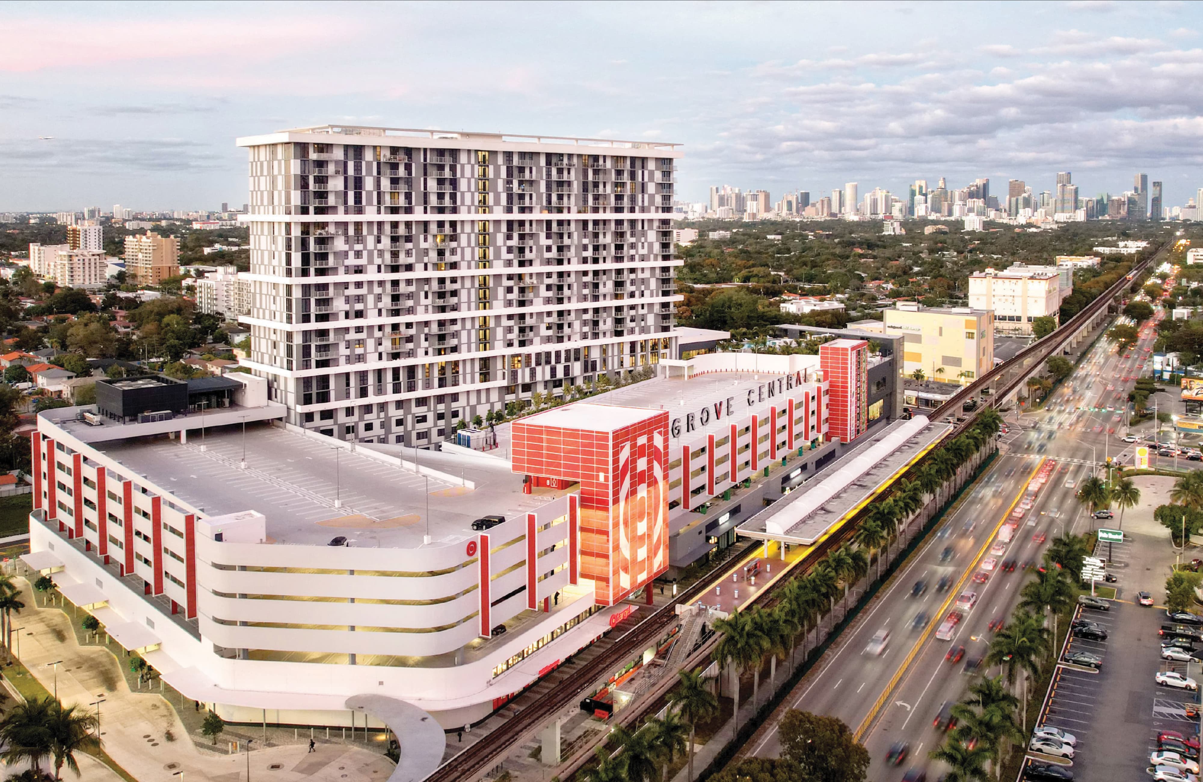 Aerial view of the Grove Central mixed-use development, showing a high-rise residential tower above a red and white retail podium and parking structure, situated alongside the Metrorail line and a busy highway, with the Miami skyline visible in the distance.