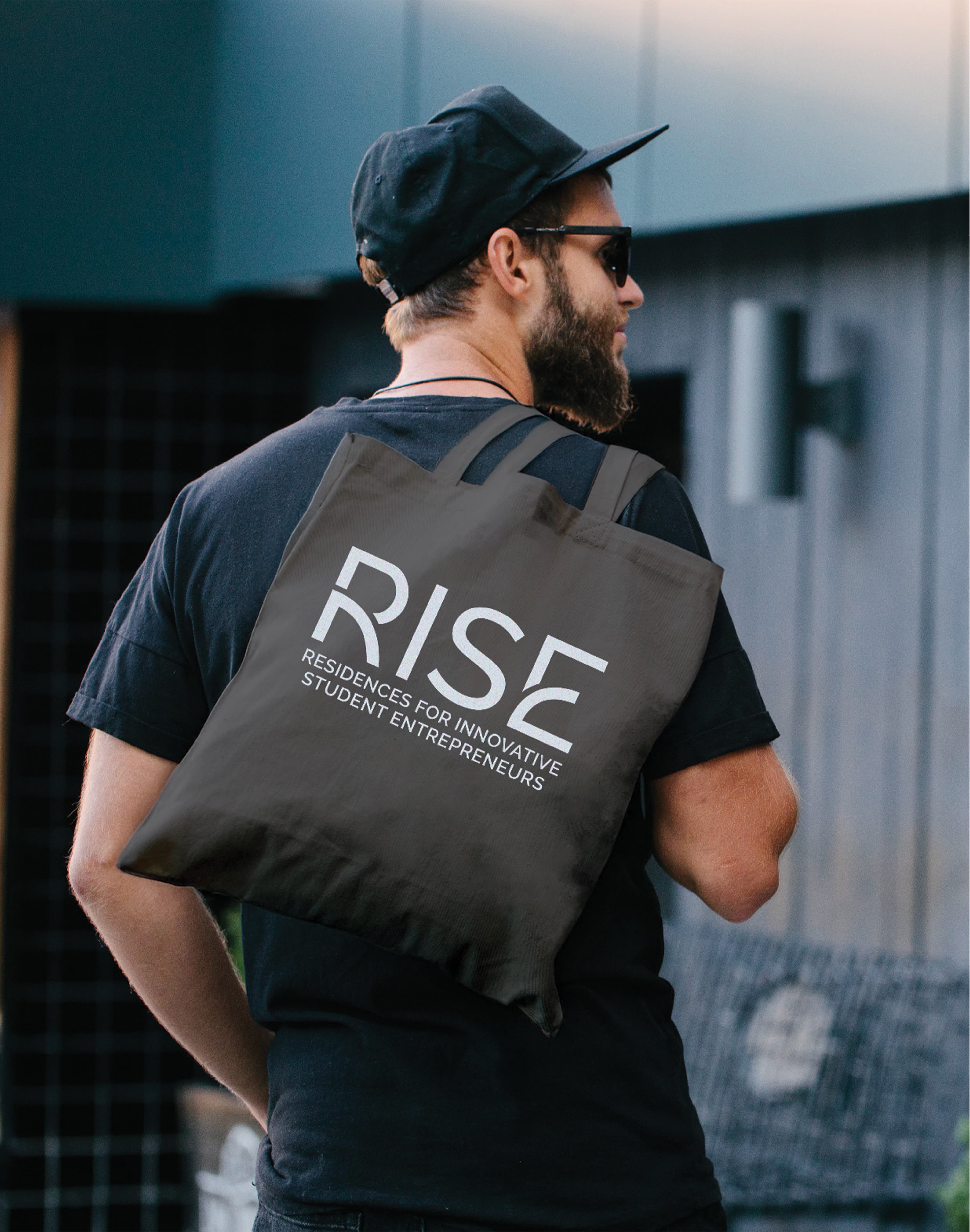 Man wearing a black cap, sunglasses, and beard seen from behind, carrying a gray tote bag over his shoulder printed with 'RISE, Residences for Innovative Student Entrepreneurs.'