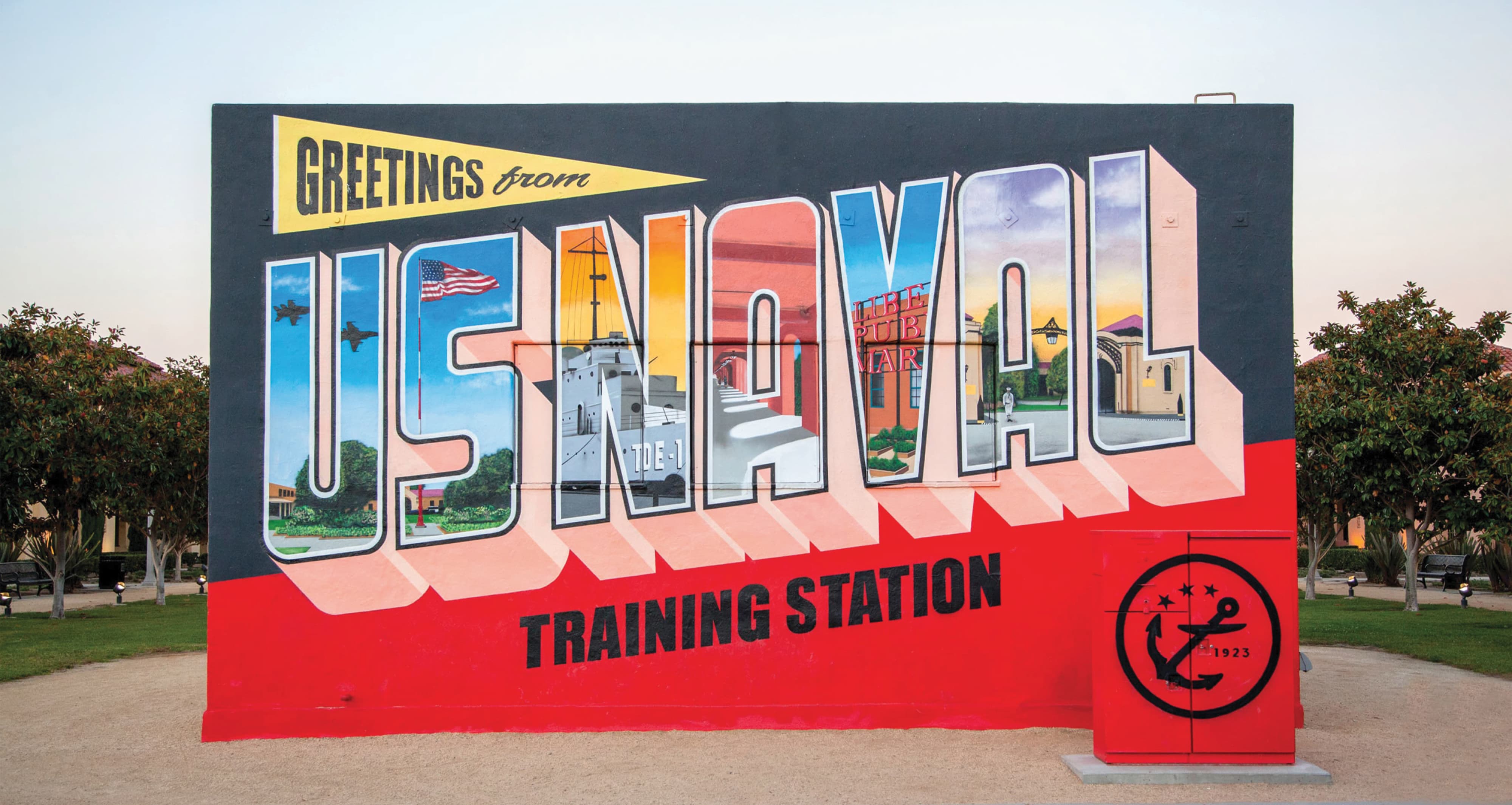 Large vintage-postcard-style mural reading 'Greetings from US Naval Training Station,' with each large letter filled with photo scenes of the base including jets, a ship, Liberty Public Market, and historic buildings, beside a red utility box stamped with the anchor and stars emblem.