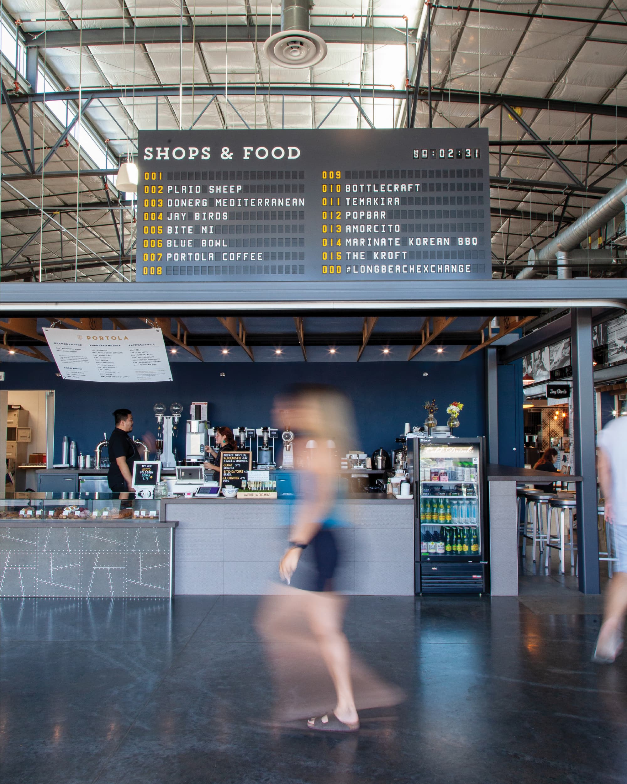 Split-flap departure board style directory reading 'Shops & Food' listing vendors like Plaid Sheep, DönerG Mediterranean, Jay Birds, and Bottlecraft, mounted above the Portola Coffee counter where baristas serve customers, as a blurred woman walks past.