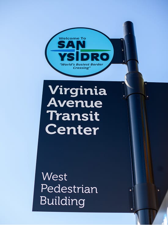 Blade sign reading 'Virginia Avenue Transit Center, West Pedestrian Building' beneath a circular 'Welcome to San Ysidro' emblem, set against a clear blue sky.