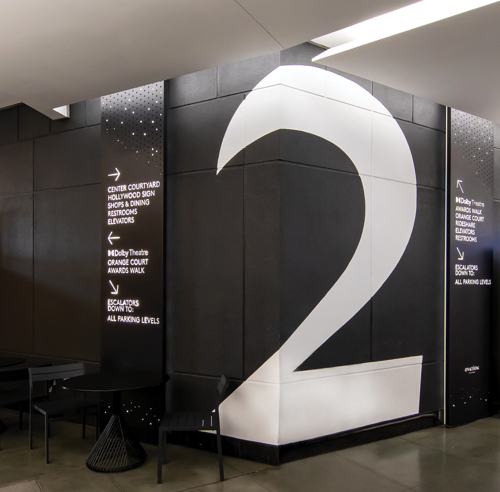 Large white level 2 painted on the corner of a black wall with vertical directional sign on each side of the number by RSM Design for Ovation Hollywood in Los Angeles, California