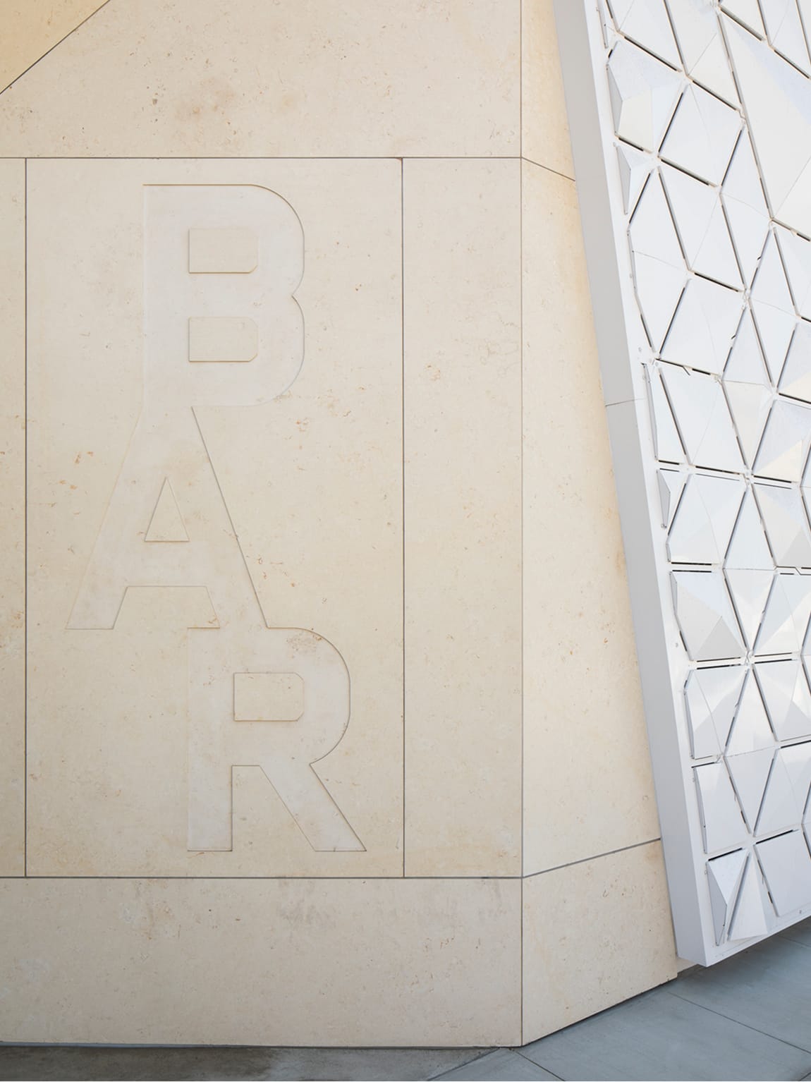 Architectural detail of the BAR Architects firm identity signage featuring large-scale tonal letterforms CNC-routed or sandblasted into a warm limestone concrete canopy wall panel, adjacent to the building's signature three-dimensional white geometric triangular facade cladding system, showcasing integrated exterior branding and material contrast