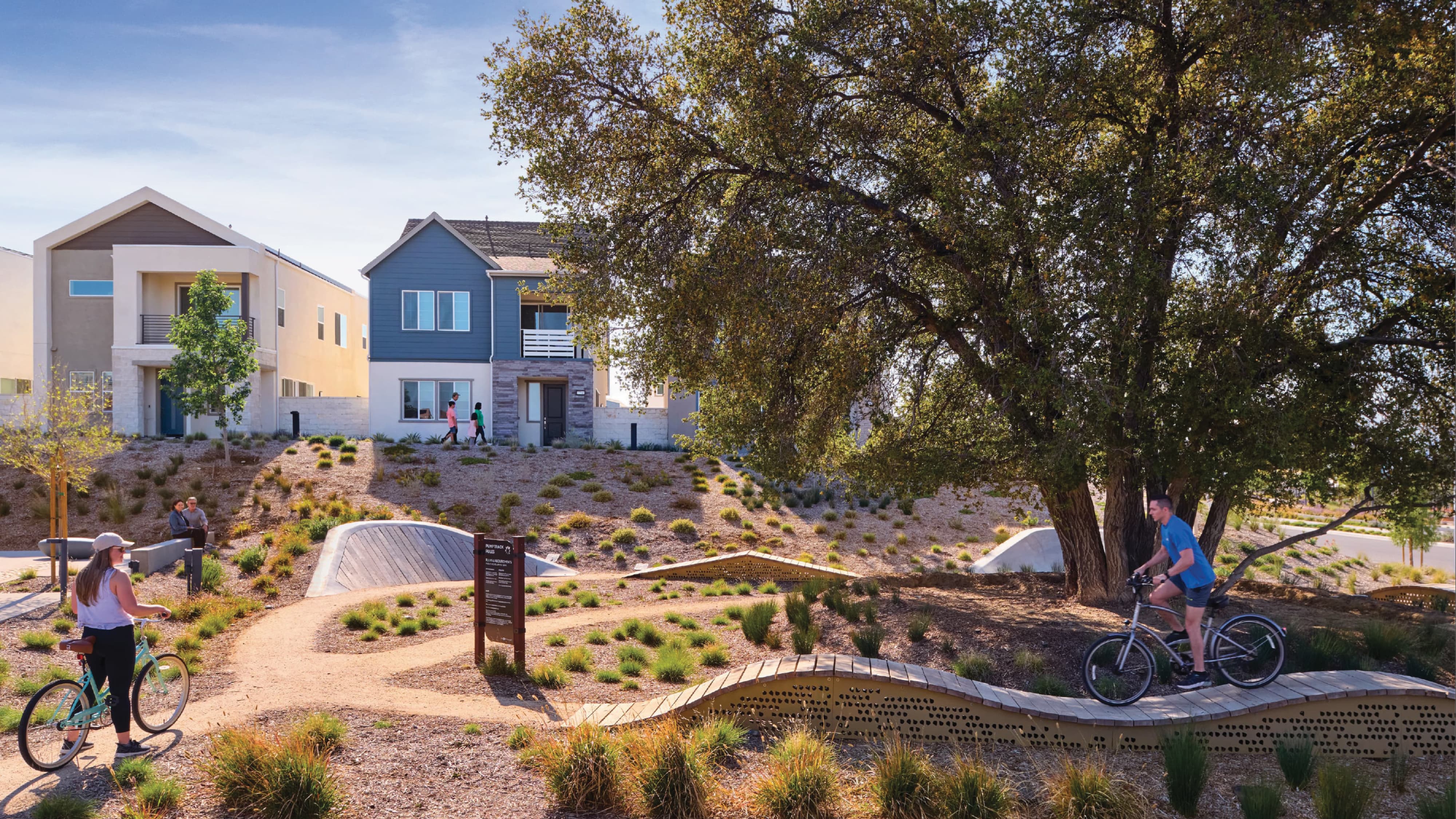 Neighborhood pump track park with people walking bikes along a curved wooden and metal track, an informational sign, and desert landscaping, with two-story homes and a large oak tree in the background.