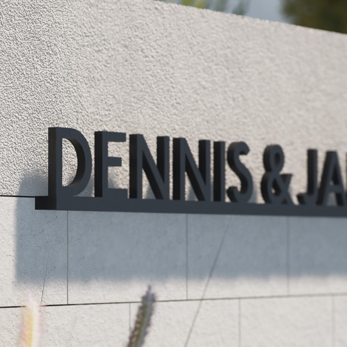 Close-up angled view of dimensional black letters reading 'Dennis & Ja...' mounted on a textured concrete block wall, with ornamental grasses partially visible in the foreground.