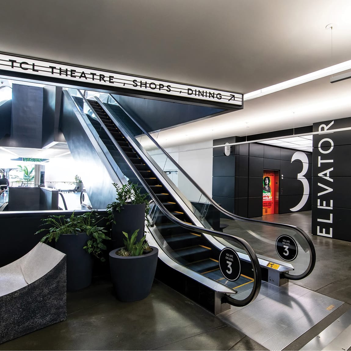 Ovation Hollywood Level 3 escalator concourse with bold black and white wayfinding signage directing to TCL Theatre, shops and dining, with large elevator number pylon and Bob Marley movie poster