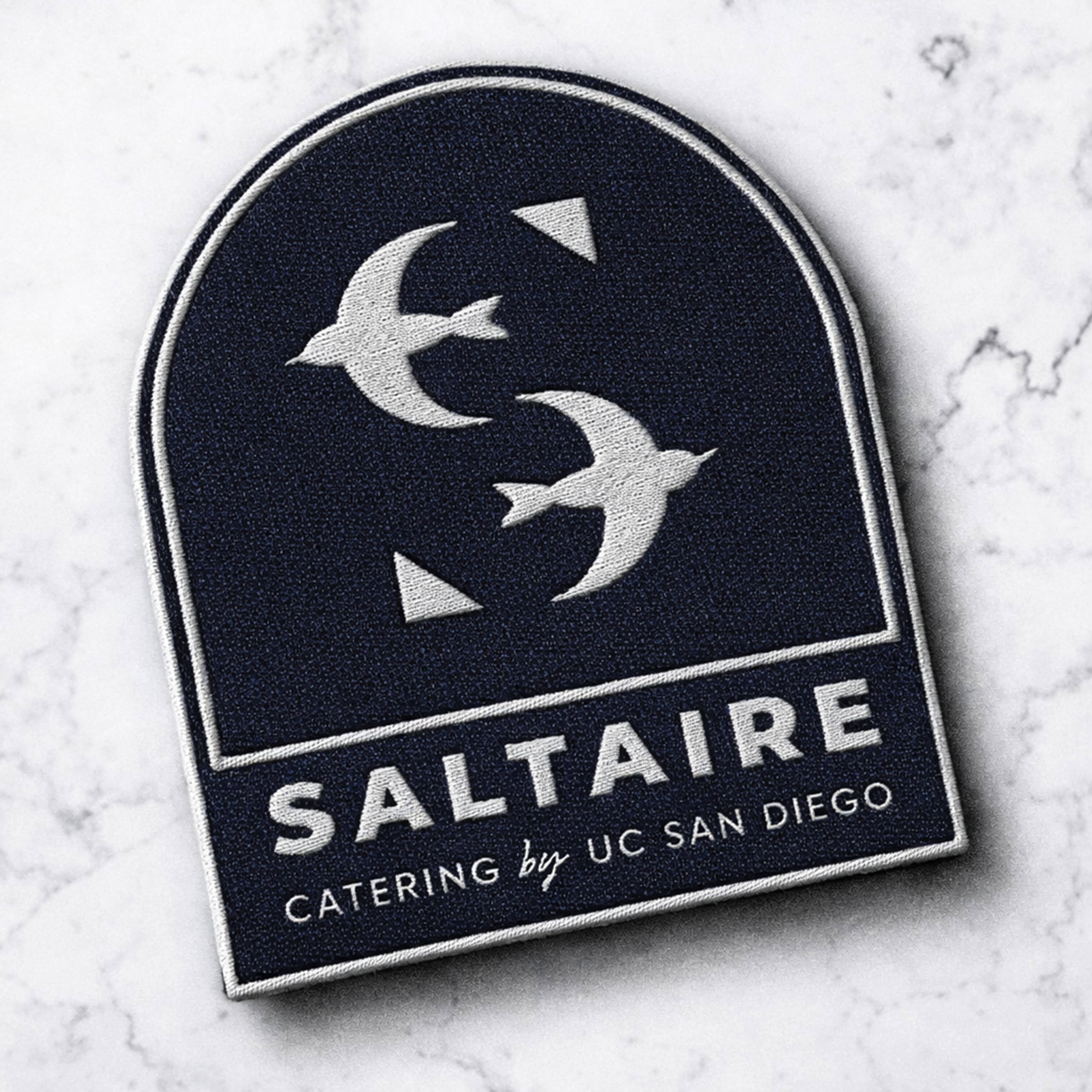 Saltaire Catering by UC San Diego embroidered badge logo featuring the tern lettermark icon in an arched navy and silver design on a marble surface, showcasing branding and visual identity design for a food and beverage and education market client by RSM Design.