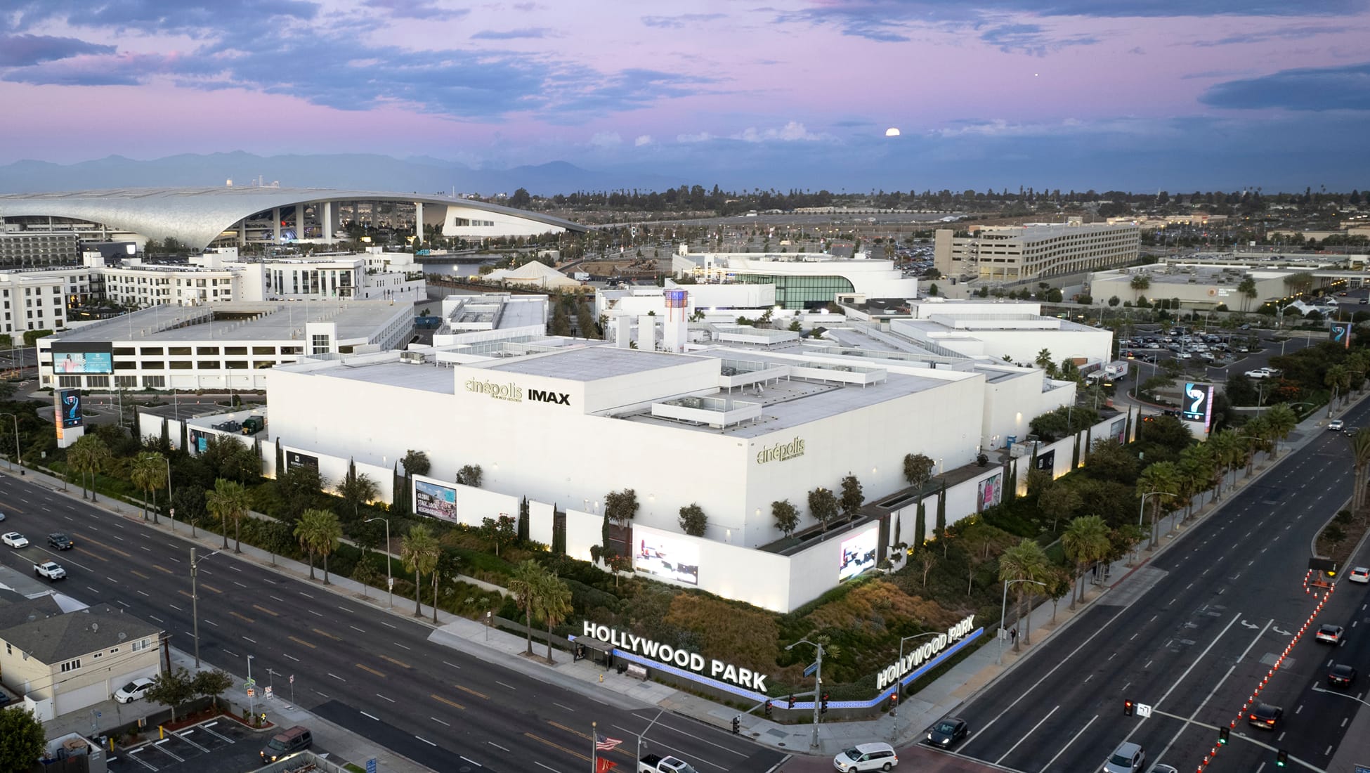 Aerial dusk view of Hollywood Park mixed-use development in Inglewood, California with Cinépolis IMAX theater, retail complex, palm tree-lined boulevards, illuminated signage, and SoFi Stadium in background