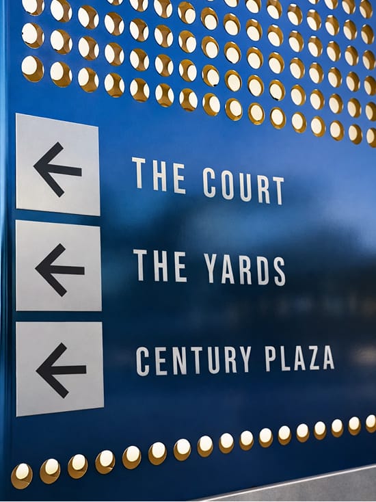 Hollywood Park interior wayfinding sign with gold dot decorative panel and directional arrows pointing to The Court, The Yards, and Century Plaza destinations, Inglewood, California
