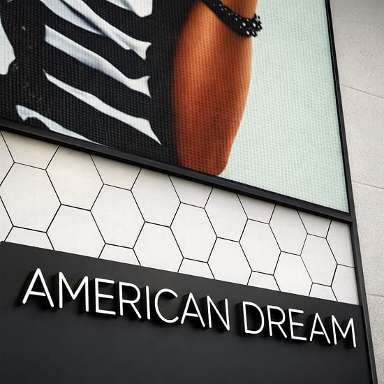 Close-up detail of the American Dream pylon sign tower base in East Rutherford, New Jersey, showing dimensional white channel letter wordmark on a matte black panel beneath a hexagonal tile graphic band and a large-scale LED fashion campaign display — showcasing RSM Design's architectural graphics, digital integration, and branded environments for a large-scale mixed-use retail and entertainment destination.
