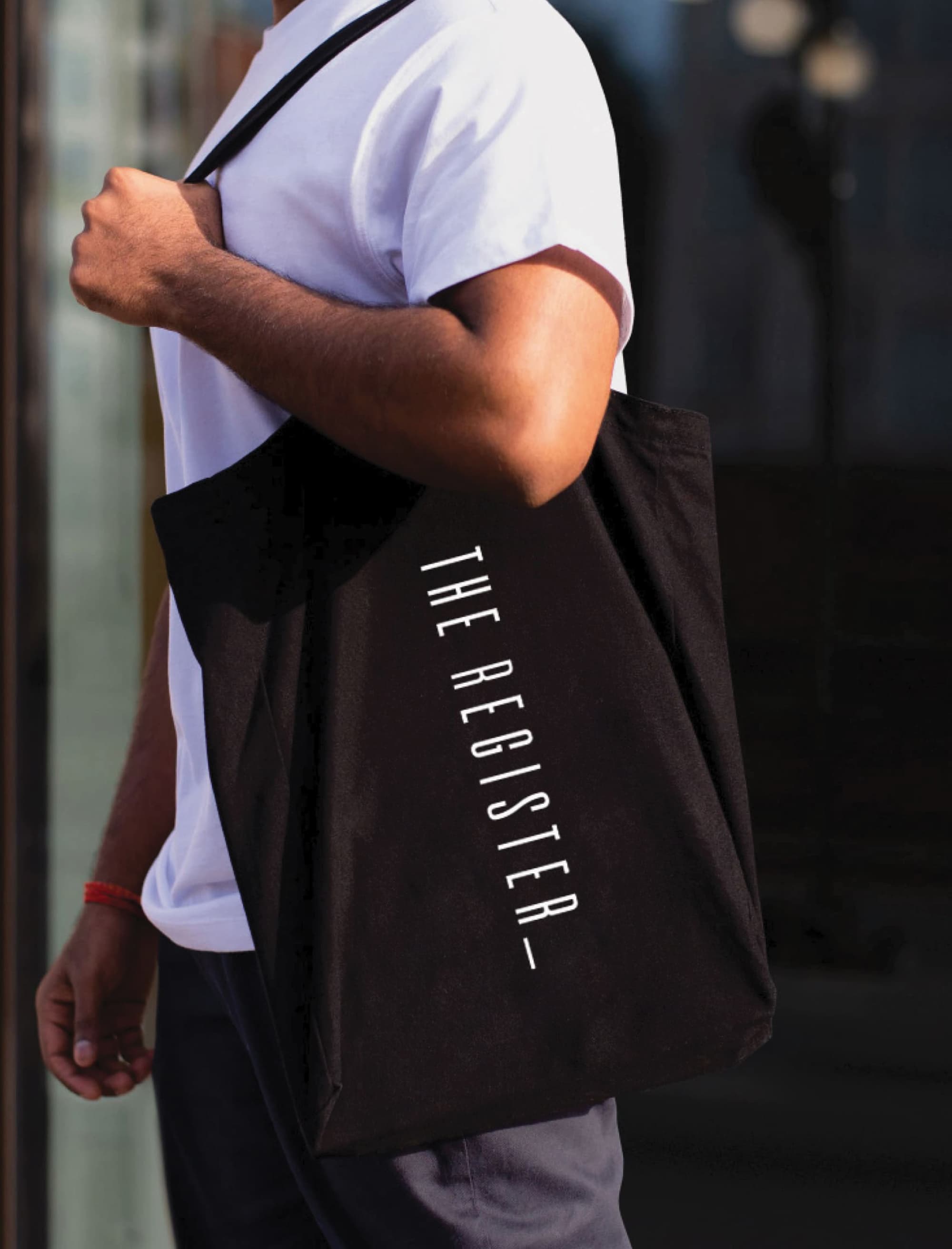 Branded black canvas tote bag with vertically oriented 'The Register' logo in white condensed typography, carried by a resident — an example of RSM Design's print and digital collateral and branding and visual identity work for a mixed-use residential community at Post District in Salt Lake City.