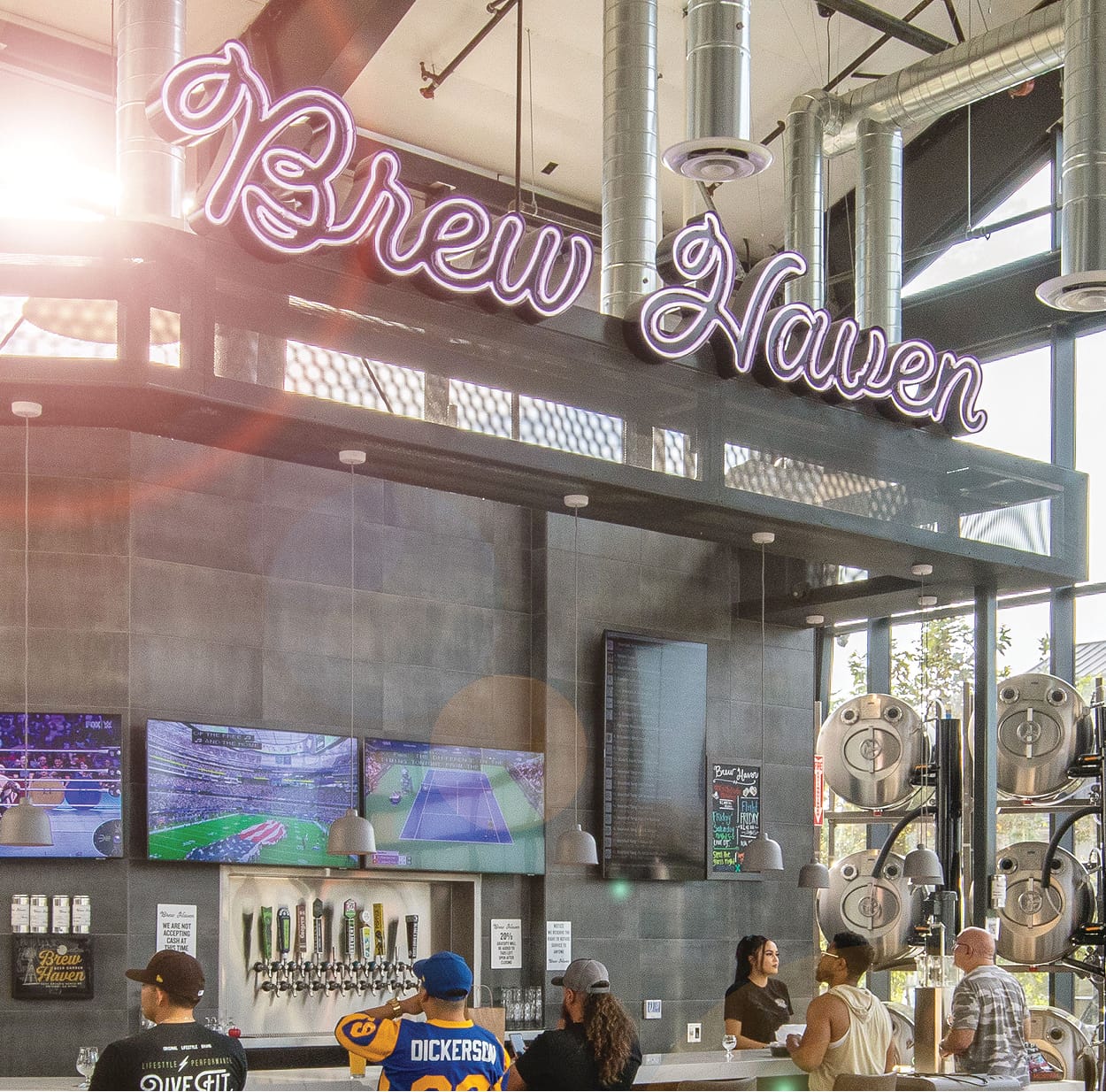 Interior of Brew Haven with a large purple neon script sign overhead, a row of beer taps and stainless steel brewing tanks behind the bar, TVs showing sports, and patrons gathered at the counter.