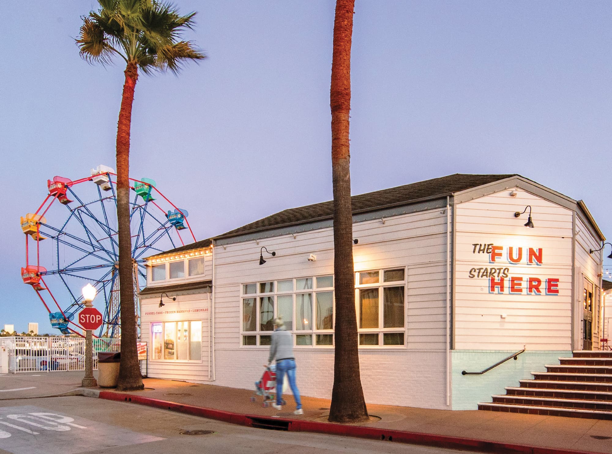 A dusk exterior view of the Balboa Fun Zone featuring a bold illuminated "The Fun Starts Here" wall sign in red and navy on a white shiplap building facade, flanked by towering palm trees, a colorful Ferris wheel, gooseneck wall sconces, and a pedestrian with a stroller passing by — showcasing RSM Design's experiential graphics and branded environments, identity signage, and placemaking services within the waterfront and mixed-use markets