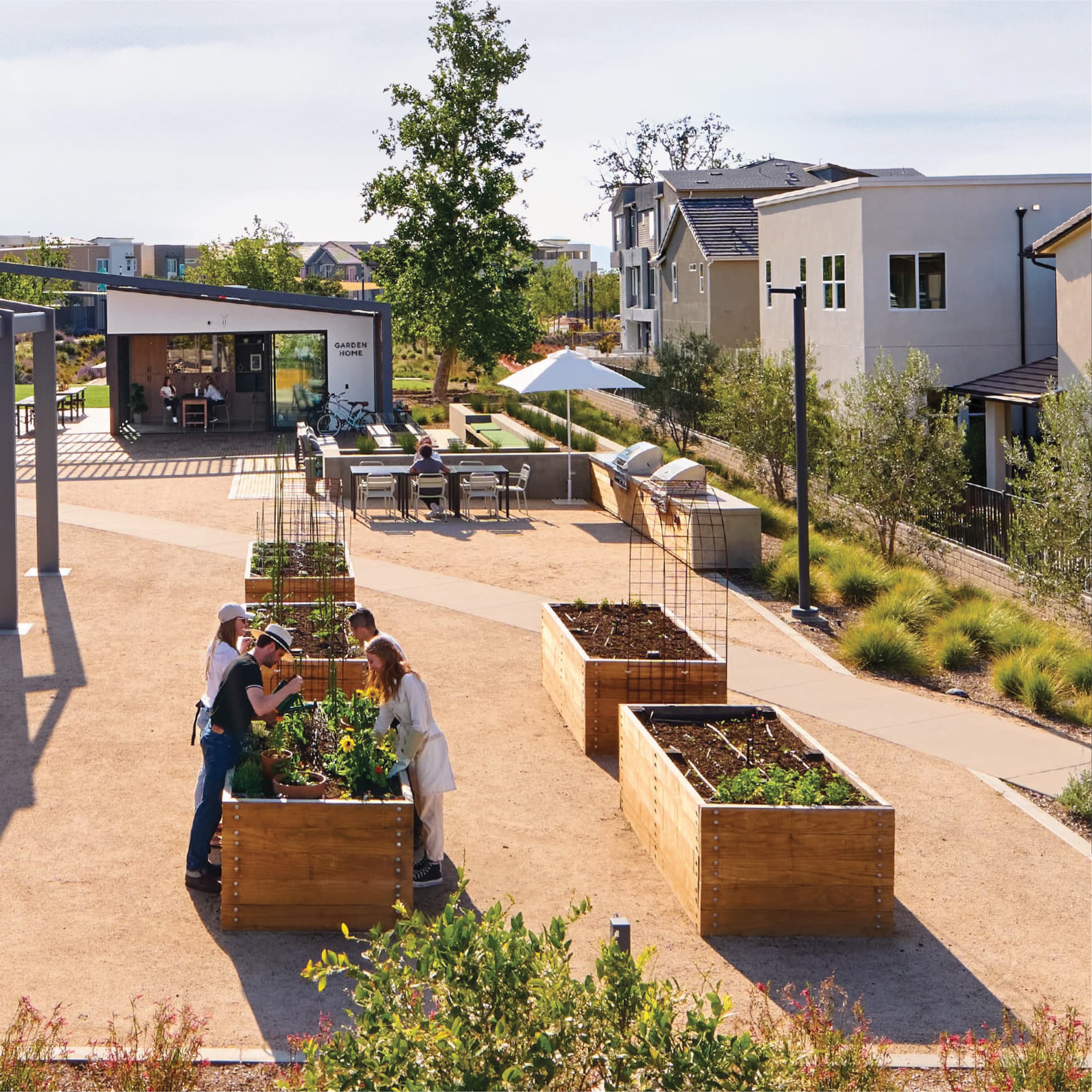 Community garden with wooden raised planter beds where people tend to flowers and vegetables, near a 'Garden Home' pavilion with outdoor seating, grills, and a shade umbrella, framed by residential buildings and native landscaping.