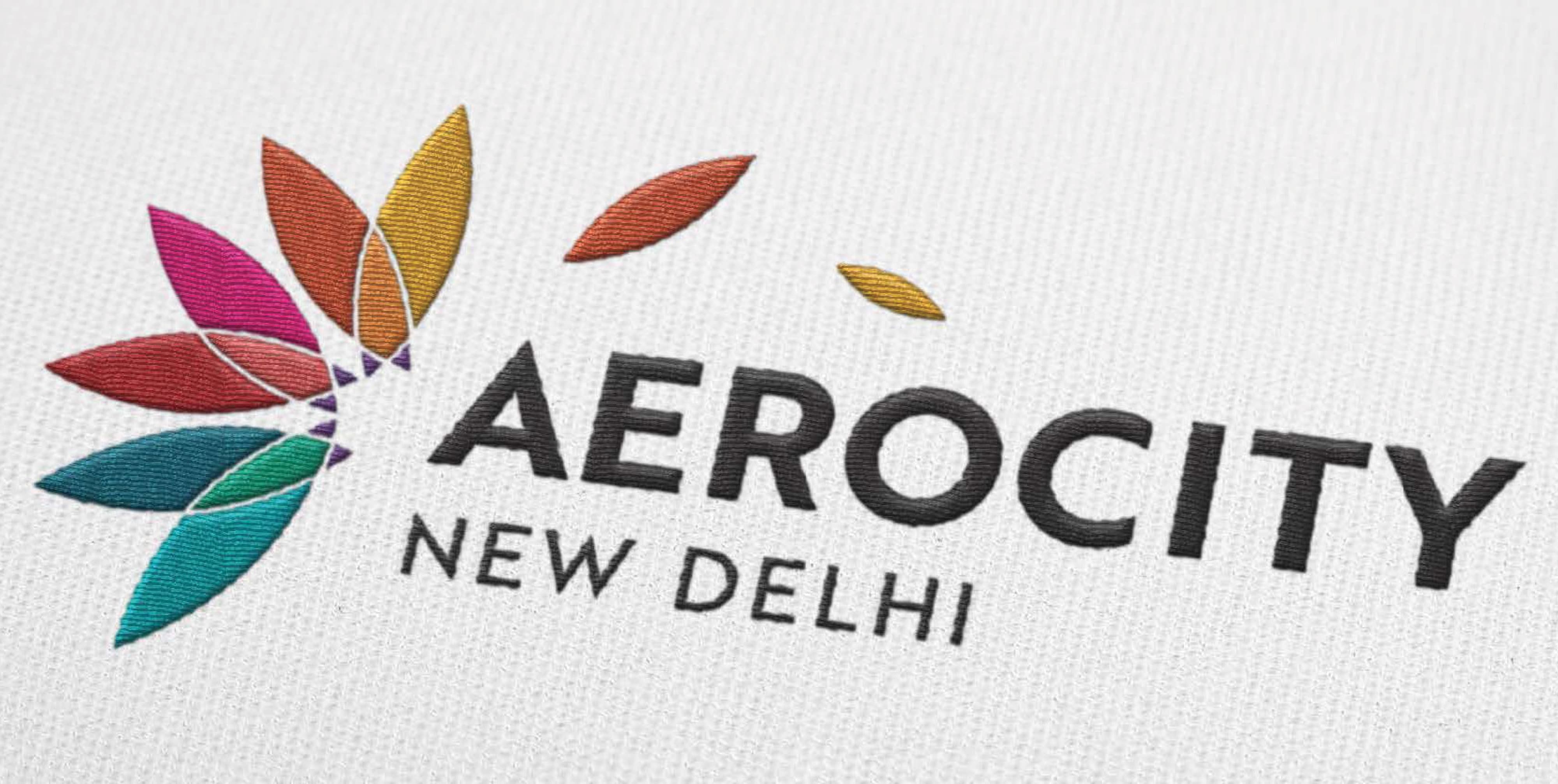 Close-up detail of Aerocity New Delhi multicolored lotus flower logo embroidered on white fabric textile merchandise, showcasing vibrant petal colors in magenta, red, orange, gold, teal, and purple — RSM Design branding and visual identity, print and digital collateral, and specialty graphics for mixed-use, transit, and international destinations.