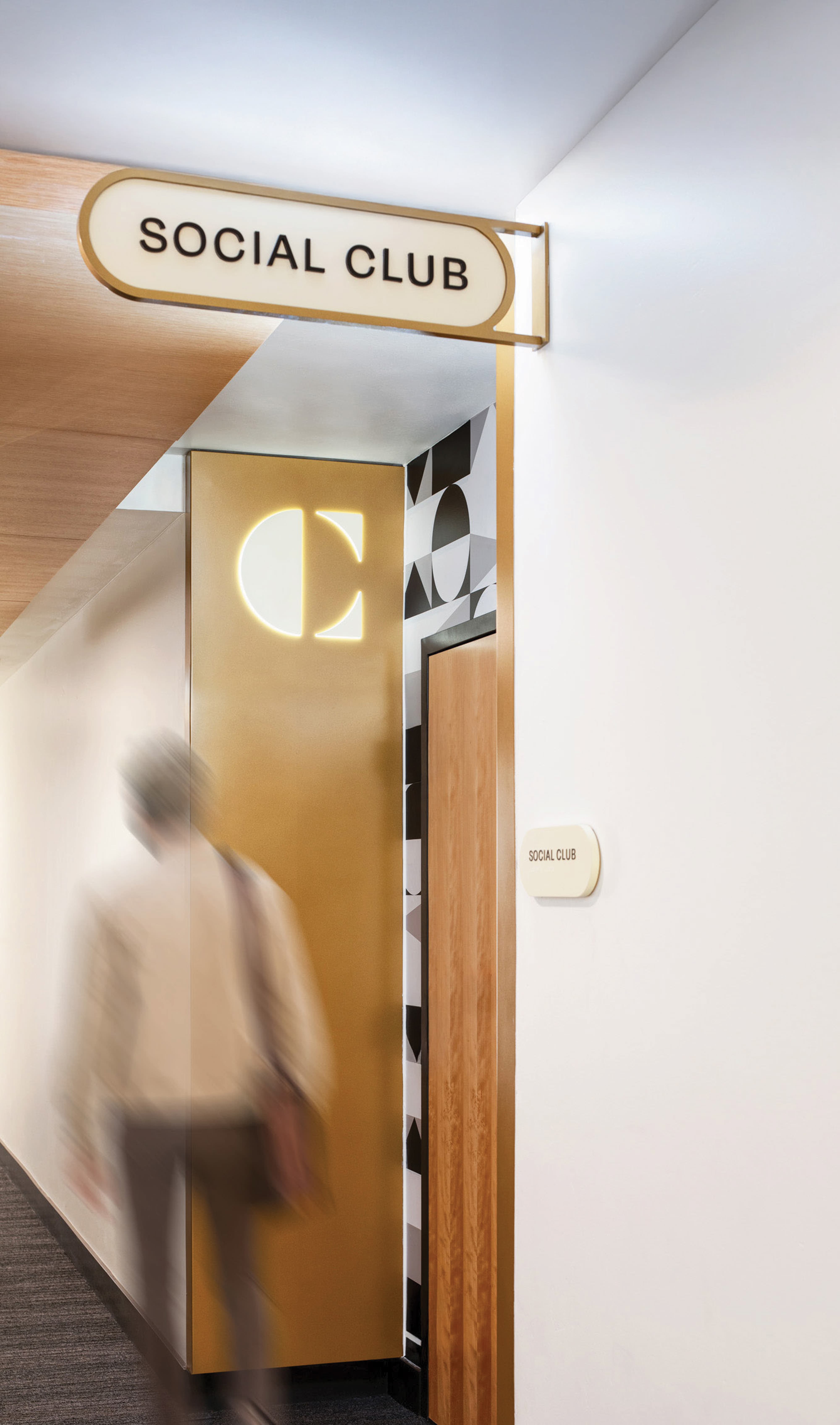 RSM Design interior wayfinding and specialty graphics for The Charles at The West Quarter in Salt Lake City, featuring a close-up of an illuminated pill-shaped overhead room identification sign and a coordinating ADA-compliant wall plaque for the Social Club amenity space, with a backlit gold panel displaying The Charles logo medallion and bold black and white geometric experiential graphics visible through the open doorway, with a motion-blurred figure entering the space, demonstrating how environmental graphic design, visual identity, and branded environments create cohesive, human-centered interior wayfinding systems for mixed-use hospitality and residential community markets.