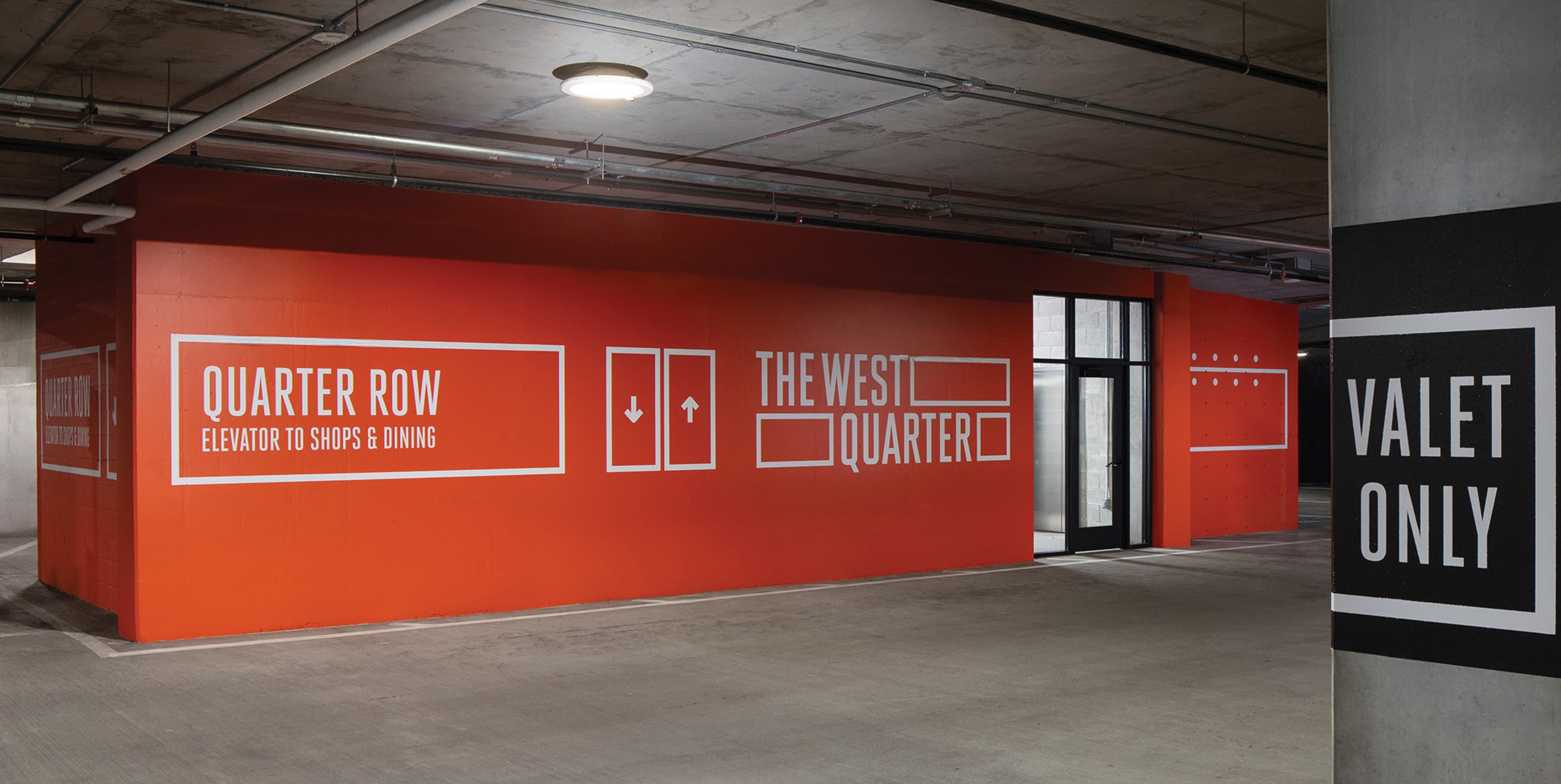 RSM Design parking garage interior wayfinding and experiential graphics for The West Quarter in Salt Lake City, featuring a bold full-wall orange mural graphic system with large-scale white typography directing visitors to Quarter Row elevator access to shops and dining, The West Quarter logo identity, elevator directional icons, and a contrasting black and white Valet Only column wrap sign, demonstrating how environmental graphic design, wayfinding and signage design, specialty graphics, and branded environments unify navigation, identity, and placemaking across every surface of a parking garage environment within a mixed-use, retail, food and beverage, residential community, and hospitality development.
