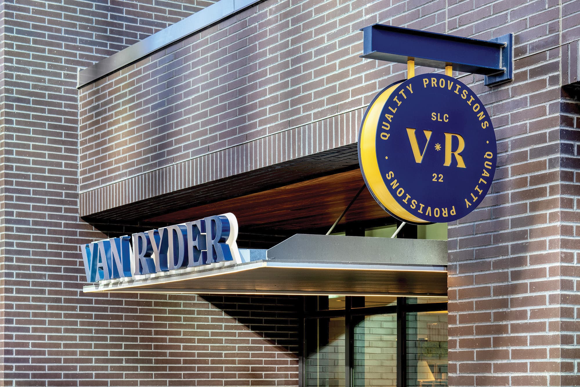 RSM Design tenant storefront signage and visual identity for Van Ryder Quality Provisions at The West Quarter in Salt Lake City, featuring dimensional navy blue channel letters mounted above a wood and metal canopy entry alongside a projecting circular medallion blade sign with gold VR monogram and circular typographic lockup on a navy background, integrated into a brick facade, showcasing how specialty graphics, environmental graphic design, master sign program design, and branded environments bring a cohesive, character-rich food and beverage identity to life within a mixed-use, hospitality, and urban streetscape market.