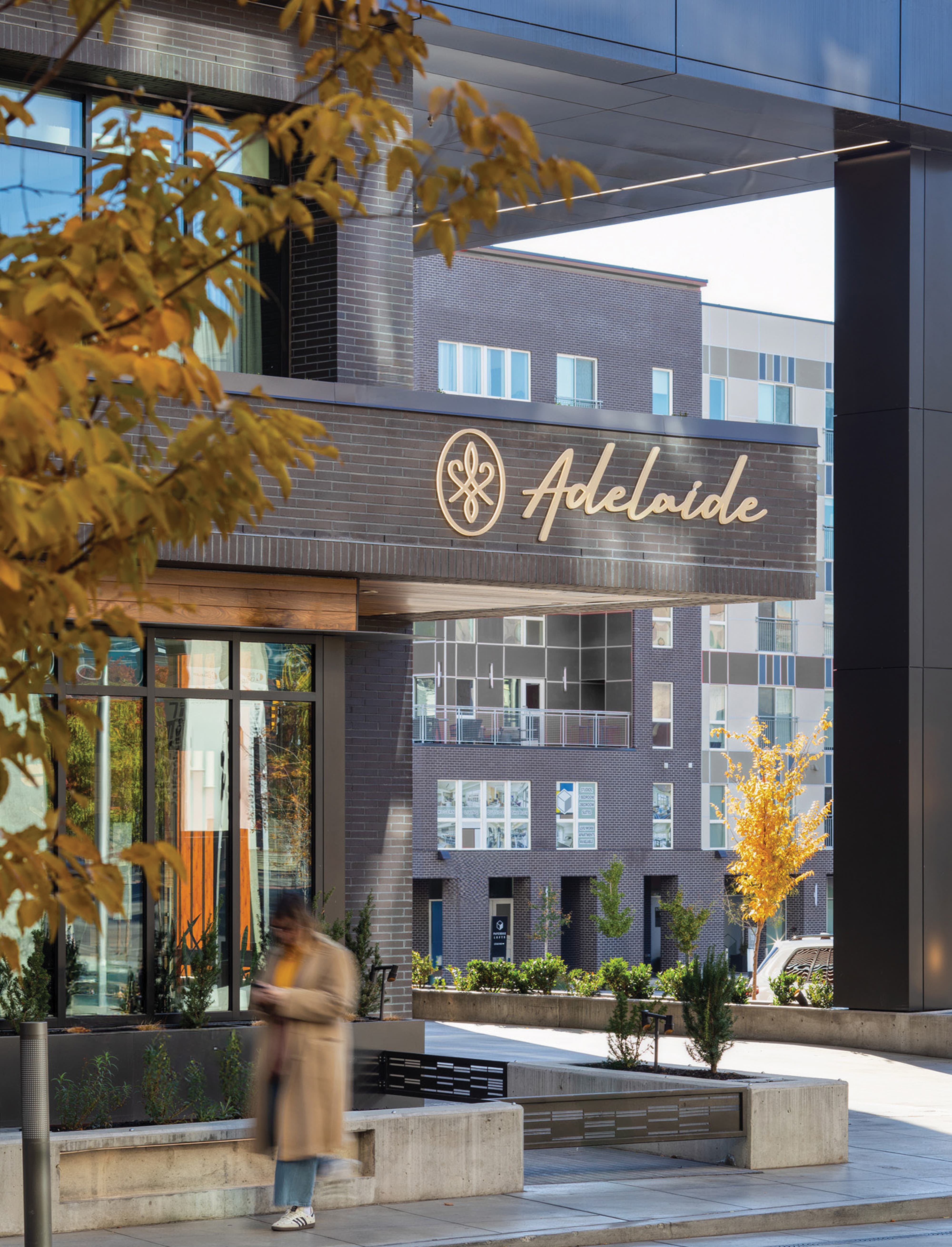 RSM Design storefront entry signage and placemaking for Adelaide restaurant at The West Quarter in Salt Lake City, featuring dimensional gold script lettering and an ornamental medallion logo set beneath a dark canopy overhang framed by a bold architectural portal, with autumn foliage in the foreground and a residential tower visible in the background, demonstrating how environmental graphic design, visual identity, and wayfinding and signage design create a layered, human-centered sense of arrival and place within a mixed-use hospitality, food and beverage, residential community, and urban streetscape development.