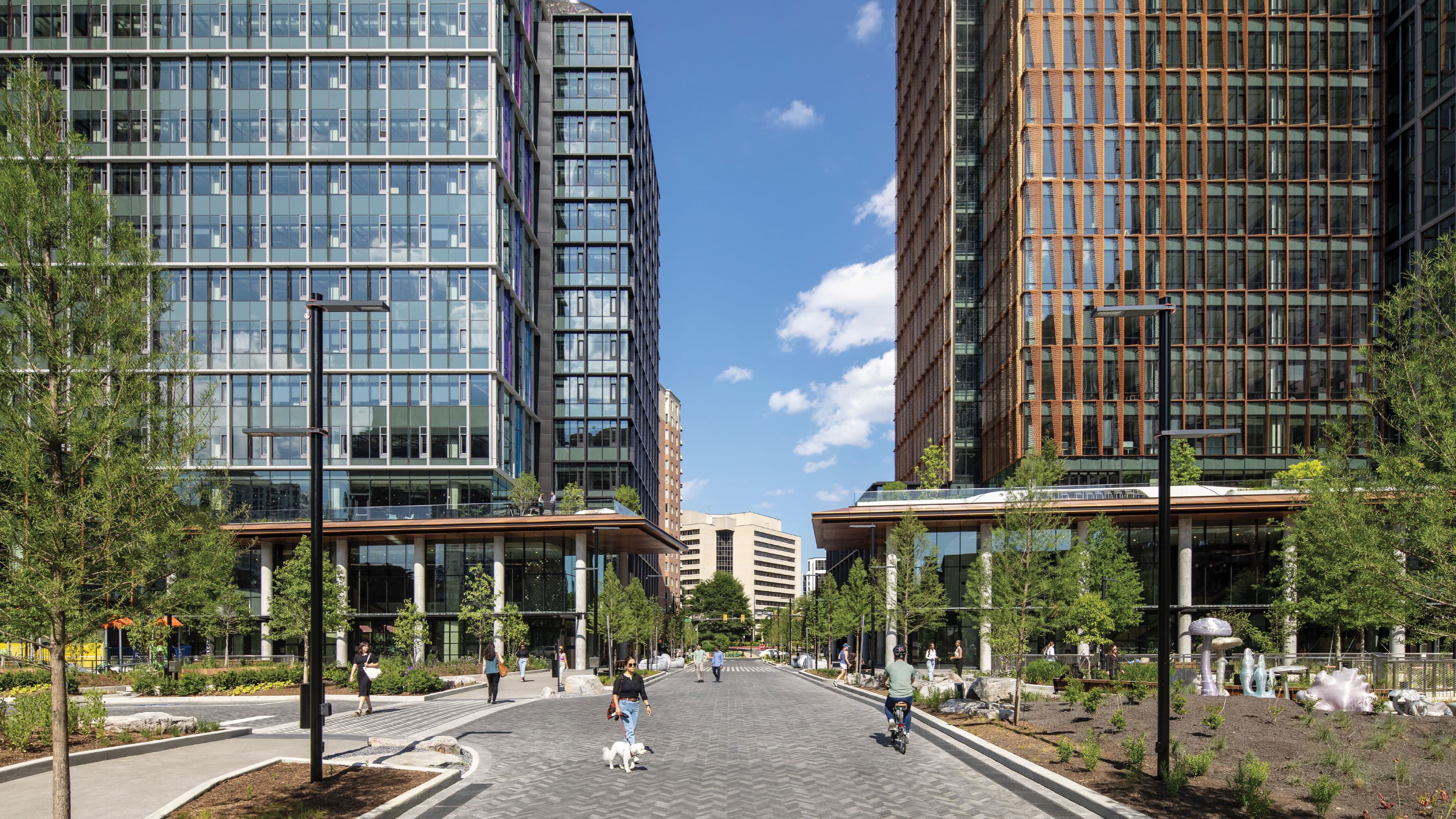 This image showcases a modern urban park area flanked by two tall, glass-covered office buildings. The scene captures a wide pedestrian-friendly pathway, landscaped with trees, plants, and contemporary sculptures. People are seen walking, biking, and engaging in leisure activities, indicating a vibrant and accessible public space. The architecture features sleek, modern design elements, including extensive use of glass and steel, with green spaces seamlessly integrated into the urban environment. This image highlights the harmonious blend of nature and urban development, creating an inviting atmosphere for both work and recreation.