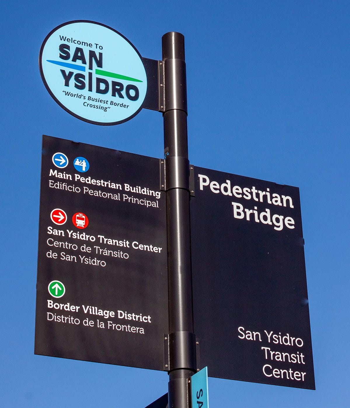 Bilingual pedestrian wayfinding sign against a blue sky, with a circular 'Welcome to San Ysidro, World's Busiest Border Crossing' emblem above, and directional panels for the Main Pedestrian Building, San Ysidro Transit Center, Border Village District, and Pedestrian Bridge.