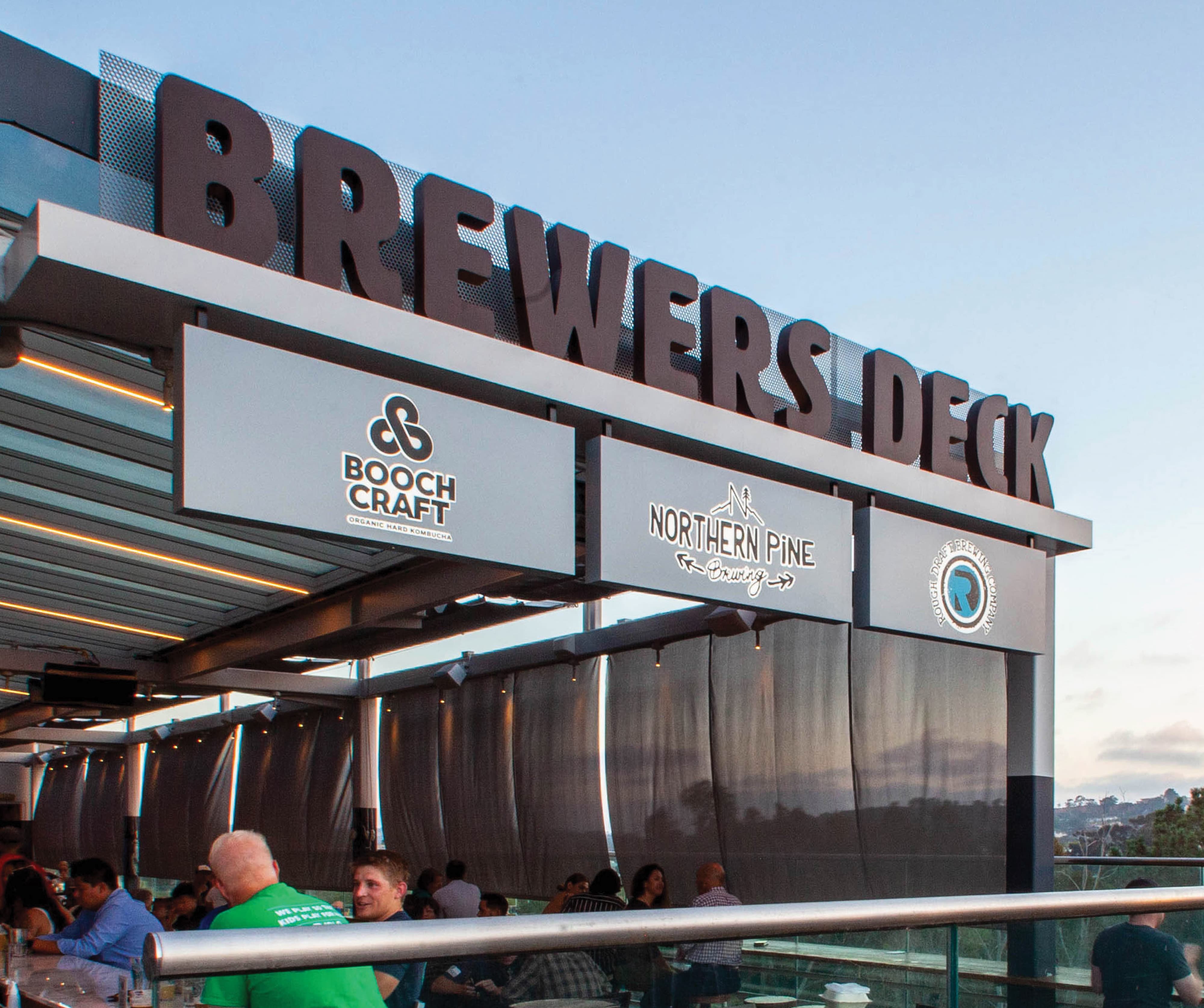 Rooftop exterior of Crafthouse Brewers Deck at dusk featuring oversized dimensional "Brewers Deck" channel letters mounted on a perforated metal facade, with illuminated tenant identity panels for Boochcraft, Northern Pine Brewing, and Rough Draft Brewing Company visible beneath a lively open-air dining terrace — showcasing RSM Design's master sign programs, tenant signage, and experiential graphics and branded environments within the food and beverage and mixed-use markets