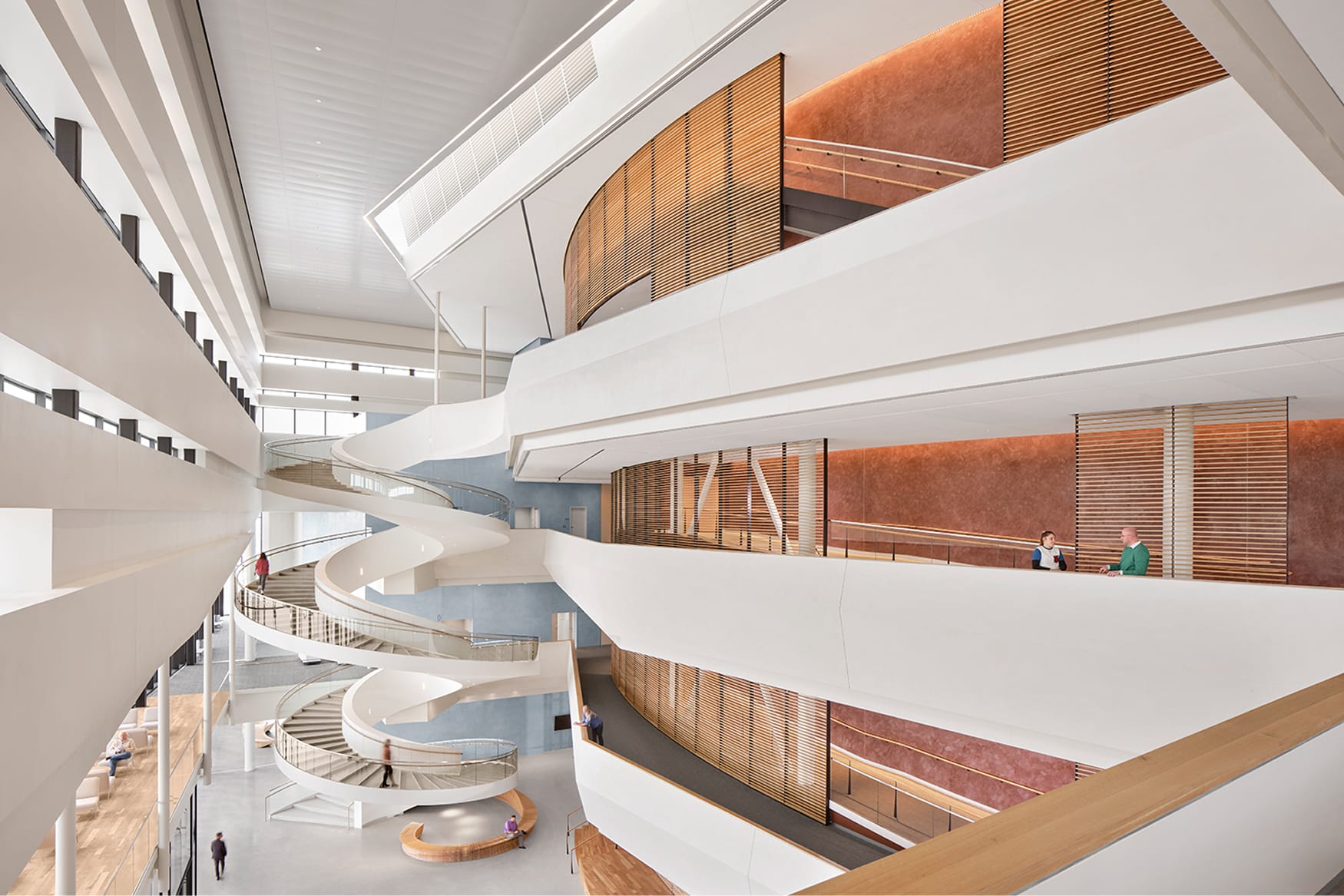 Multi-story atrium interior featuring a dramatic sculptural spiral staircase, open balconies with warm wood slat railings, terracotta accent walls, and natural light, Lubbock Independent School District administrative building