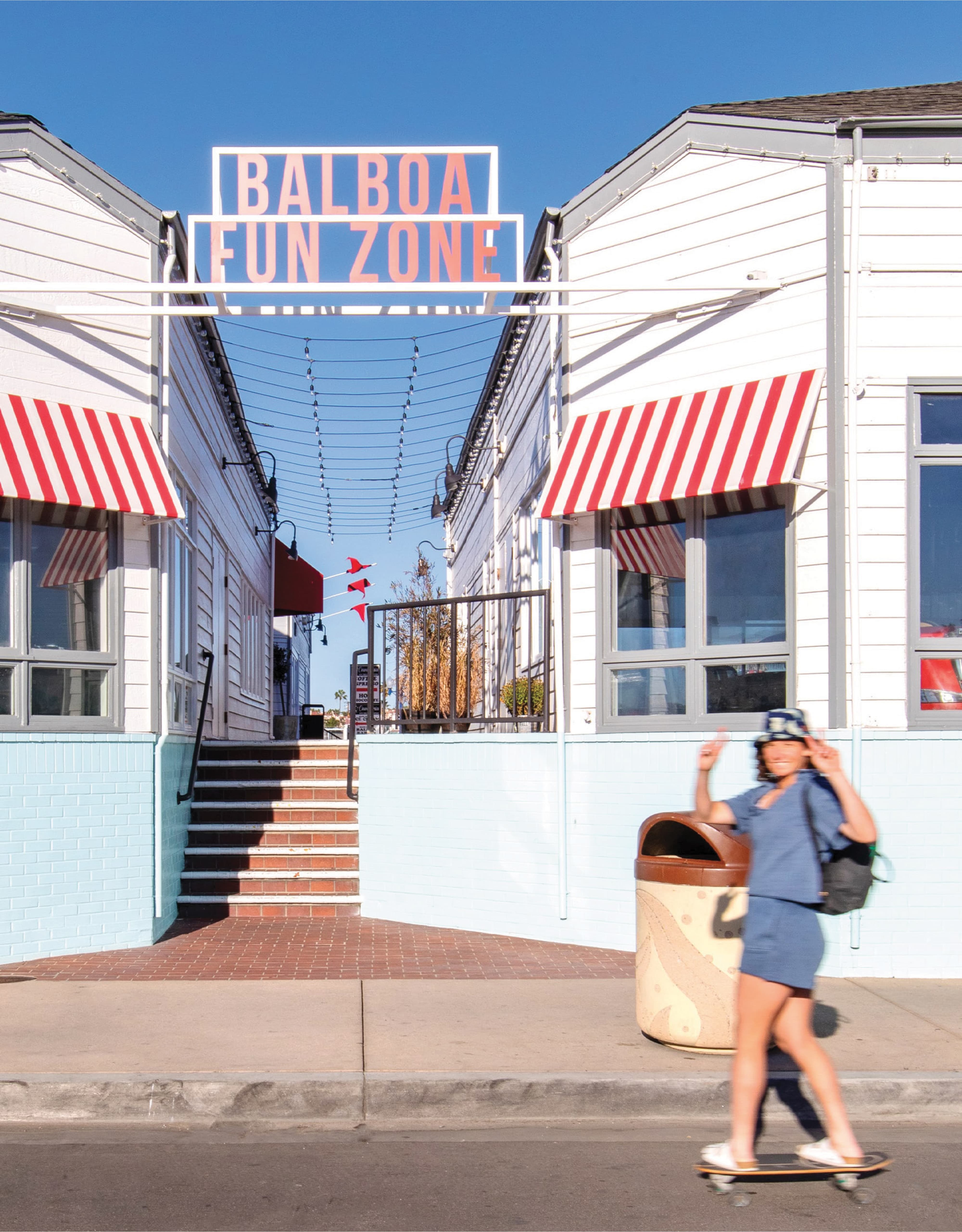 A daytime street-level view of the Balboa Fun Zone pedestrian paseo entry featuring the iconic coral and white illuminated gateway sign, red and white striped awnings, string lights overhead, a light blue brick base, brick steps leading toward the waterfront, and a skateboarder passing by in the foreground — capturing the joyful, people-centered placemaking and identity signage RSM Design delivered within the waterfront and mixed-use markets