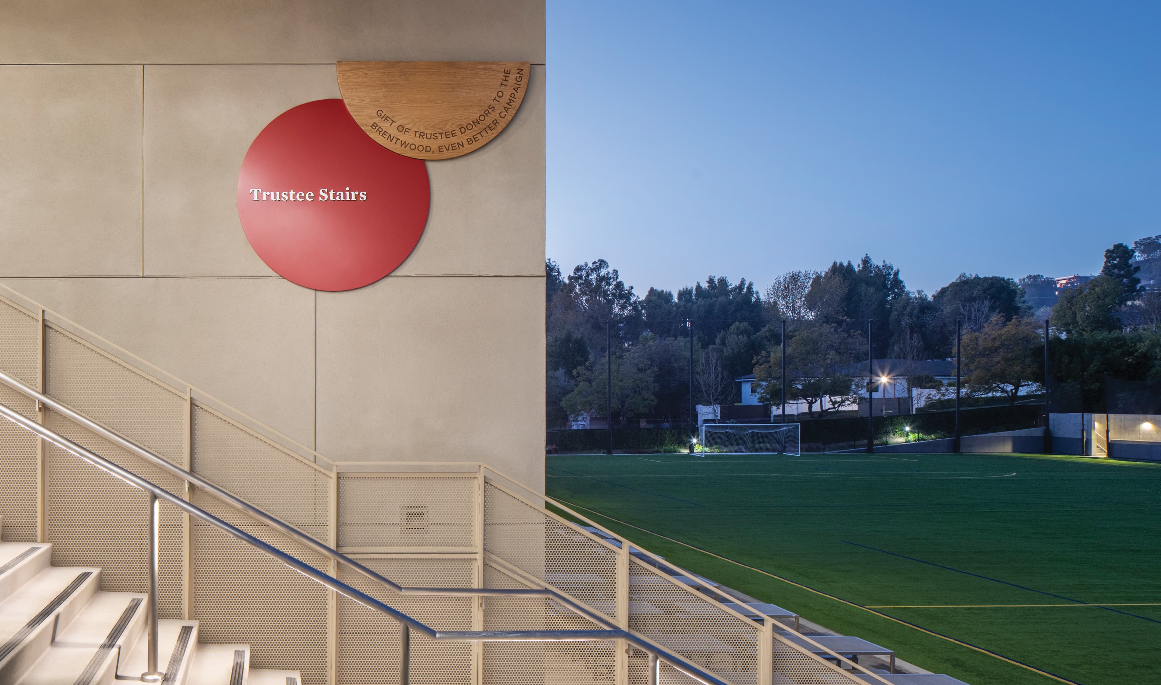 A donor recognition and space identification sign for "Trustee Stairs" composed of a red circular panel with dimensional white lettering overlapping a natural oak wood half-circle engraved with "Gift of Trustee Donors to the Brentwood, Even Better Campaign," mounted on a concrete wall alongside a perforated metal staircase overlooking an illuminated athletic field at dusk — showcasing RSM Design's donor recognition and specialty graphics and placemaking services that connect philanthropic legacy to the daily lived experience of a campus within the education market.