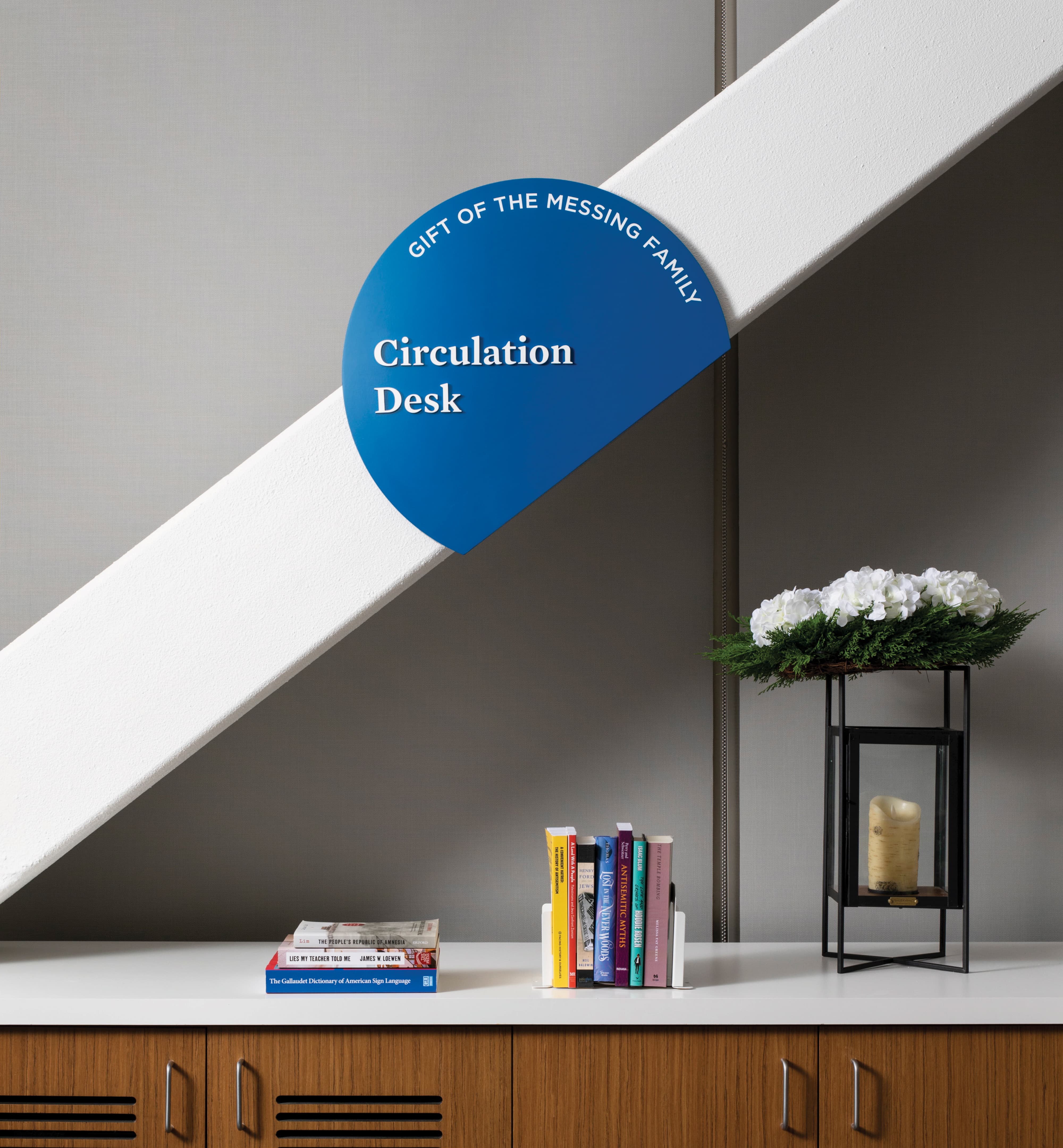 A cobalt blue circular donor recognition and space identification sign reading "Circulation Desk — Gift of the Messing Family" mounted dramatically against a white architectural staircase soffit, positioned above a library desk with a curated book display and floral arrangement — demonstrating RSM Design's donor recognition and specialty graphics services that unify wayfinding and philanthropic acknowledgment within the education market