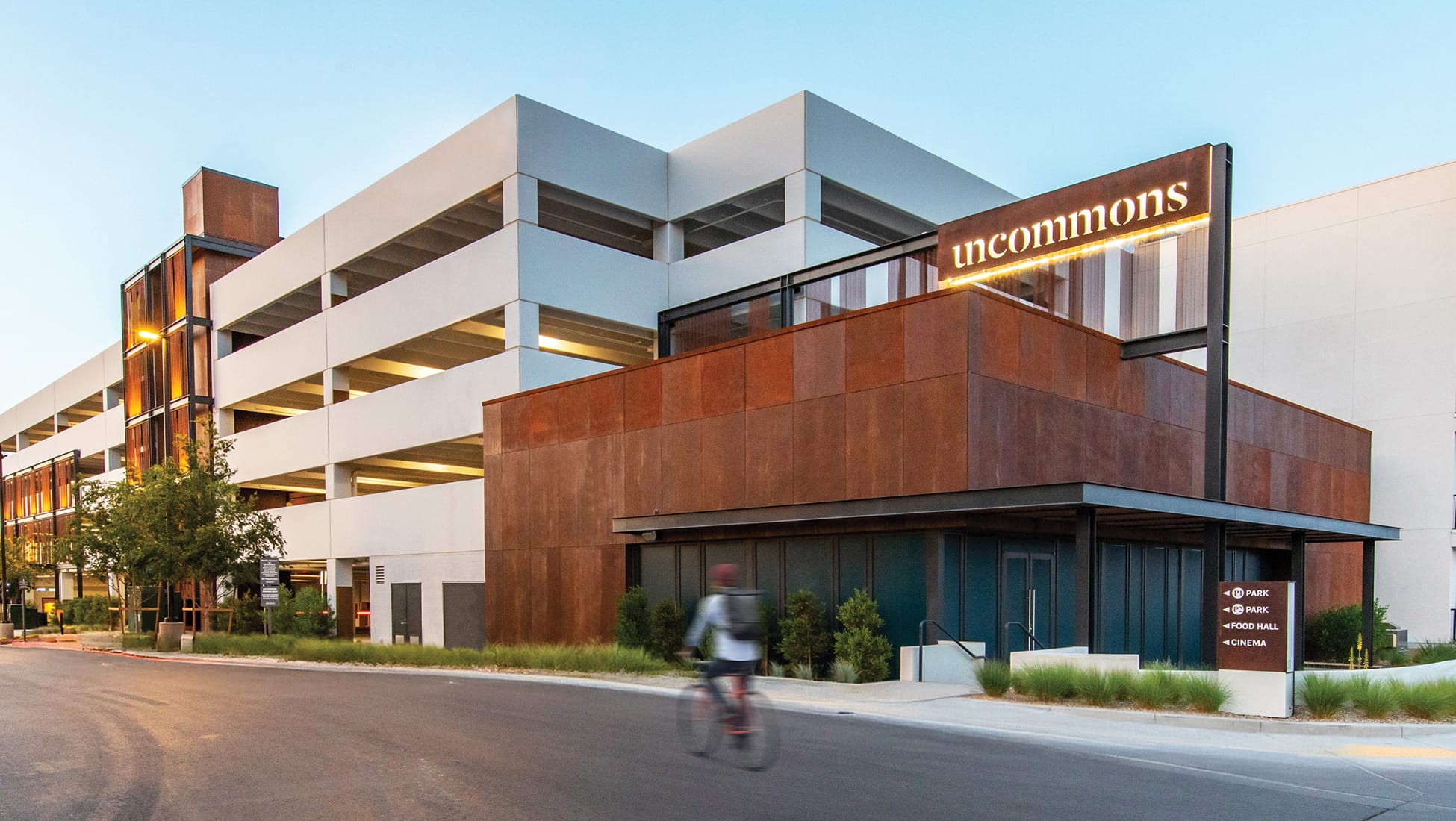Exterior view of Uncommons, a mixed-use development in Las Vegas, photographed at dusk. The building features a striking combination of weathered Corten steel cladding and white concrete cantilevered levels. An illuminated "uncommons" sign is mounted on the upper facade. A wayfinding sign near the entrance directs visitors to the park, food hall, and cinema. A cyclist passes in the foreground on the street below.
