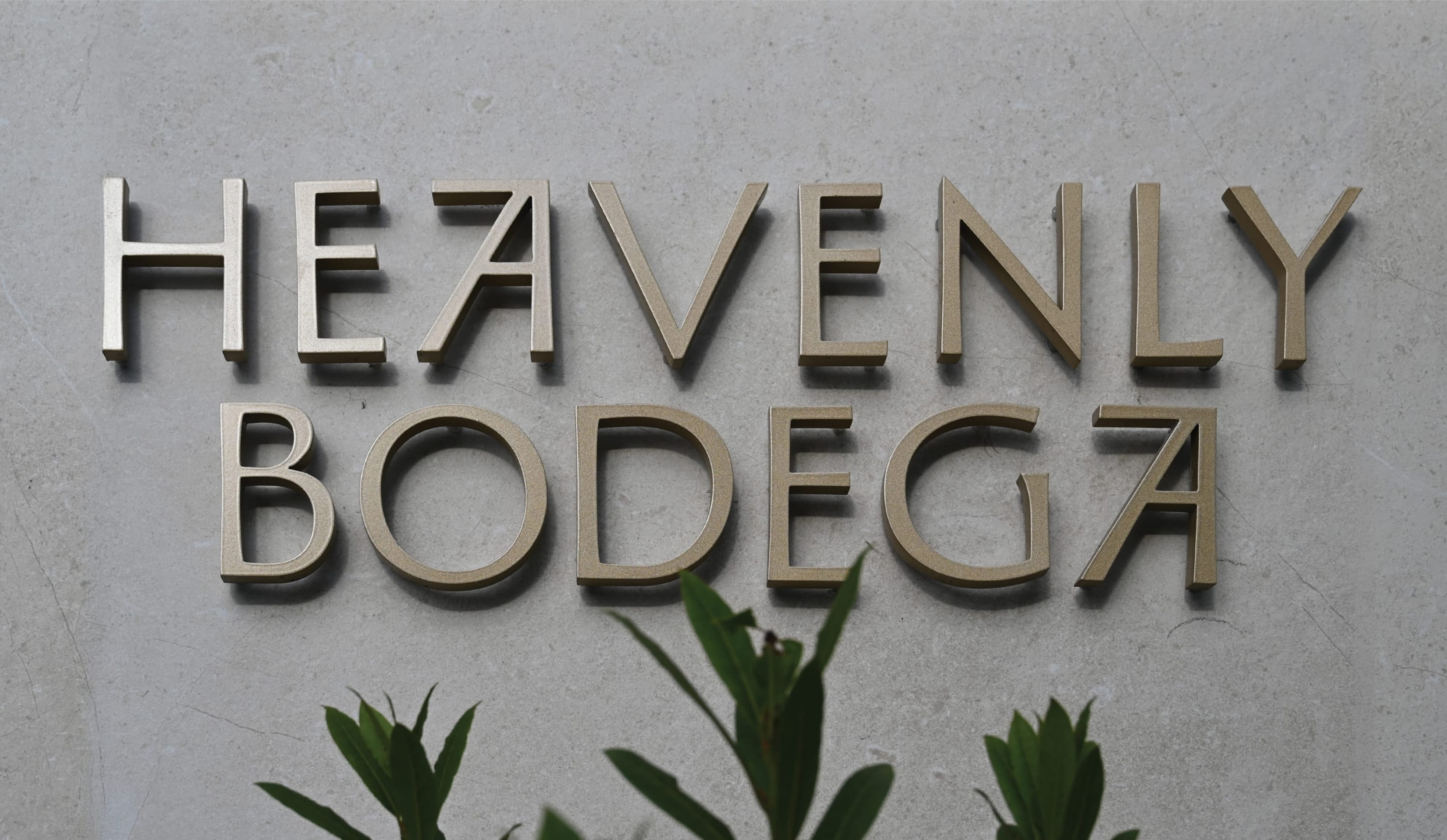 Dimensional gold "Heavenly Bodega" letters mounted on a concrete exterior wall with green foliage below — exterior signage design by RSM Design