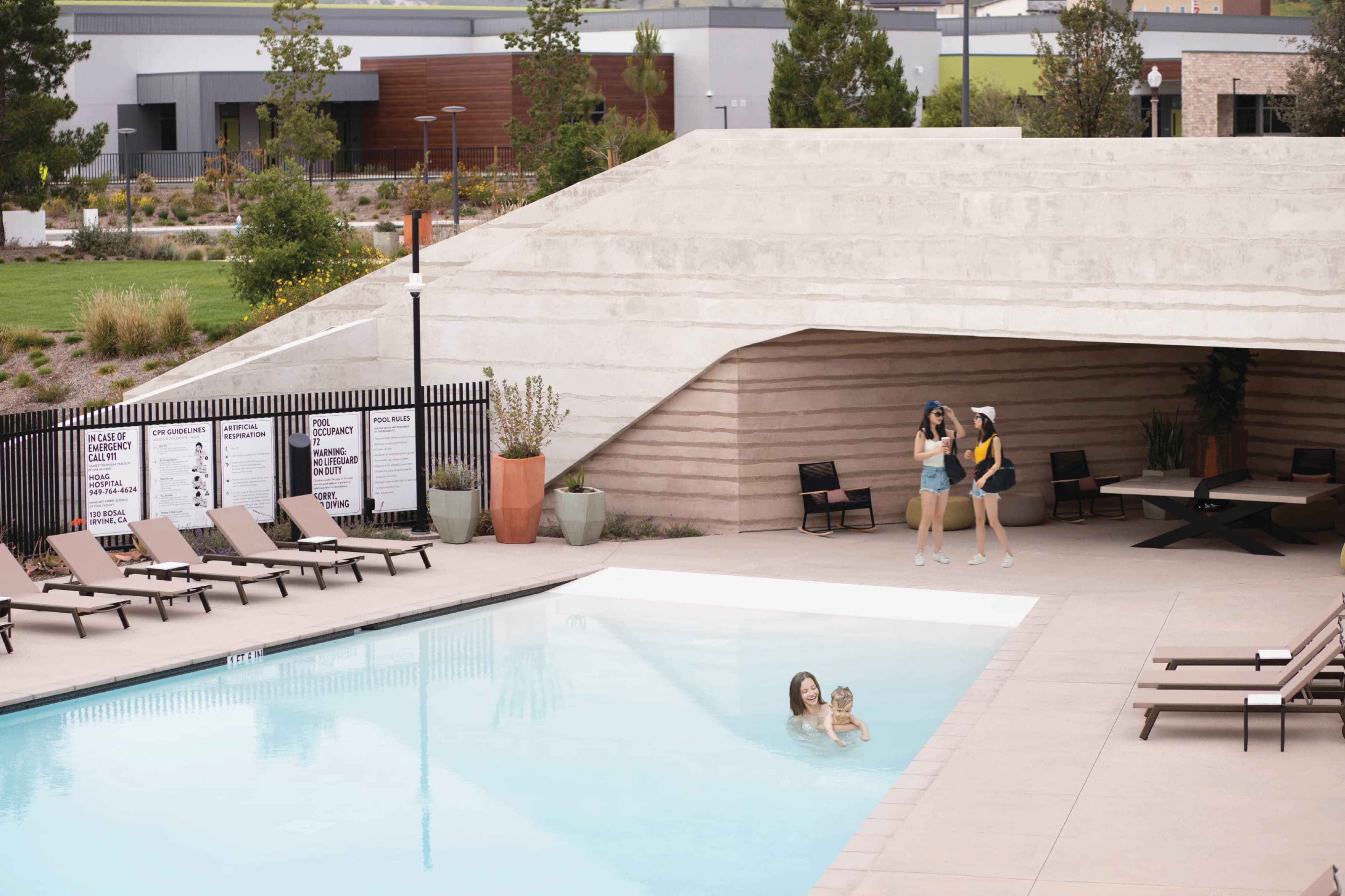 An elevated view of the Solis Park community pool amenity featuring residents swimming and socializing alongside a series of branded regulatory and safety signs — including pool rules, CPR guidelines, artificial respiration, and emergency contact information — mounted on a black perimeter fence adjacent to lounge chairs and a sculptural rammed earth shade structure — illustrating RSM Design's wayfinding and signage design and environmental graphic design services applied comprehensively across the residential community market