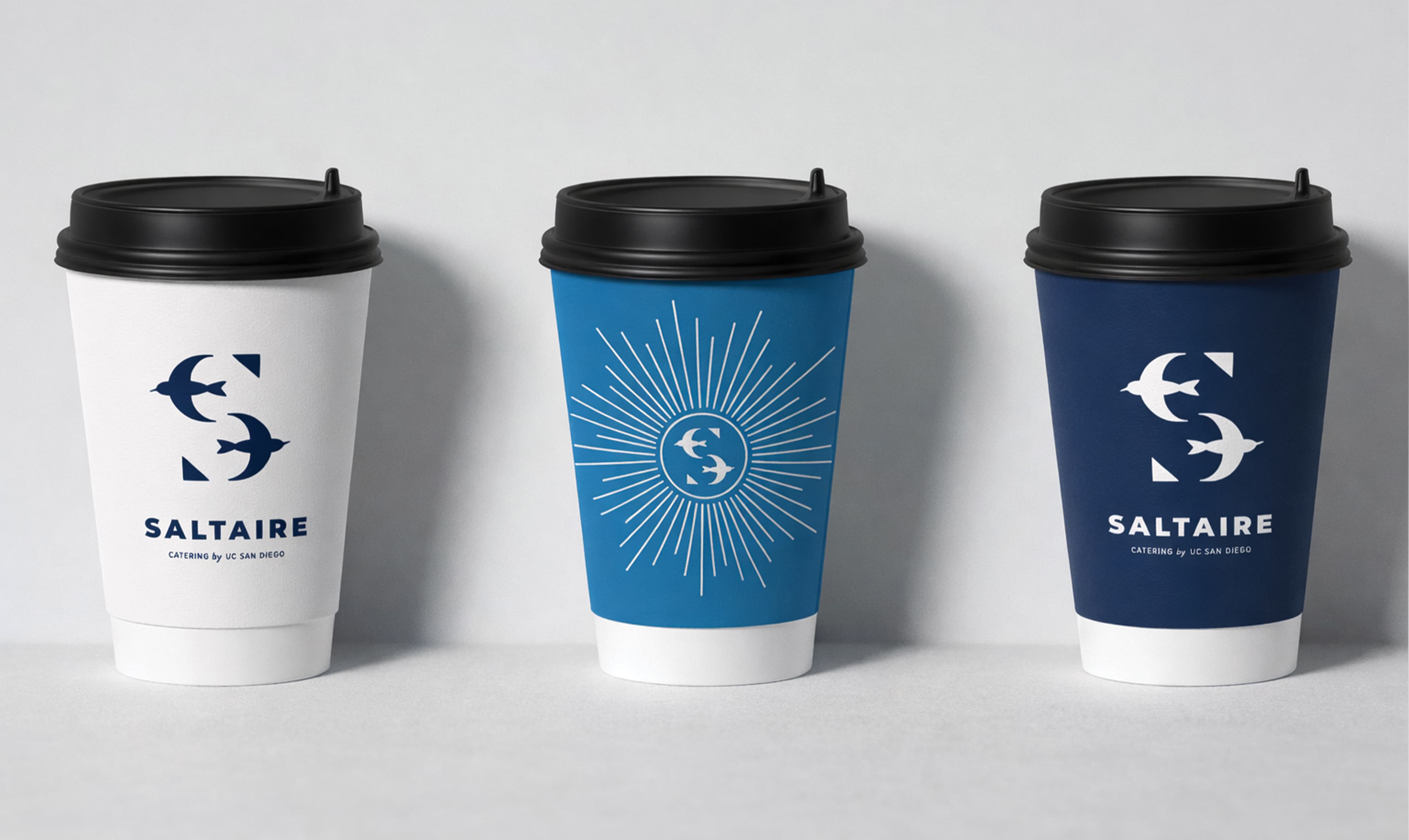 Three Saltaire Catering by UC San Diego branded coffee cups in white, ocean blue, and navy colorways featuring the tern lettermark and sunburst icon, showcasing print collateral and visual identity design for a food and beverage and education market client by RSM Design.