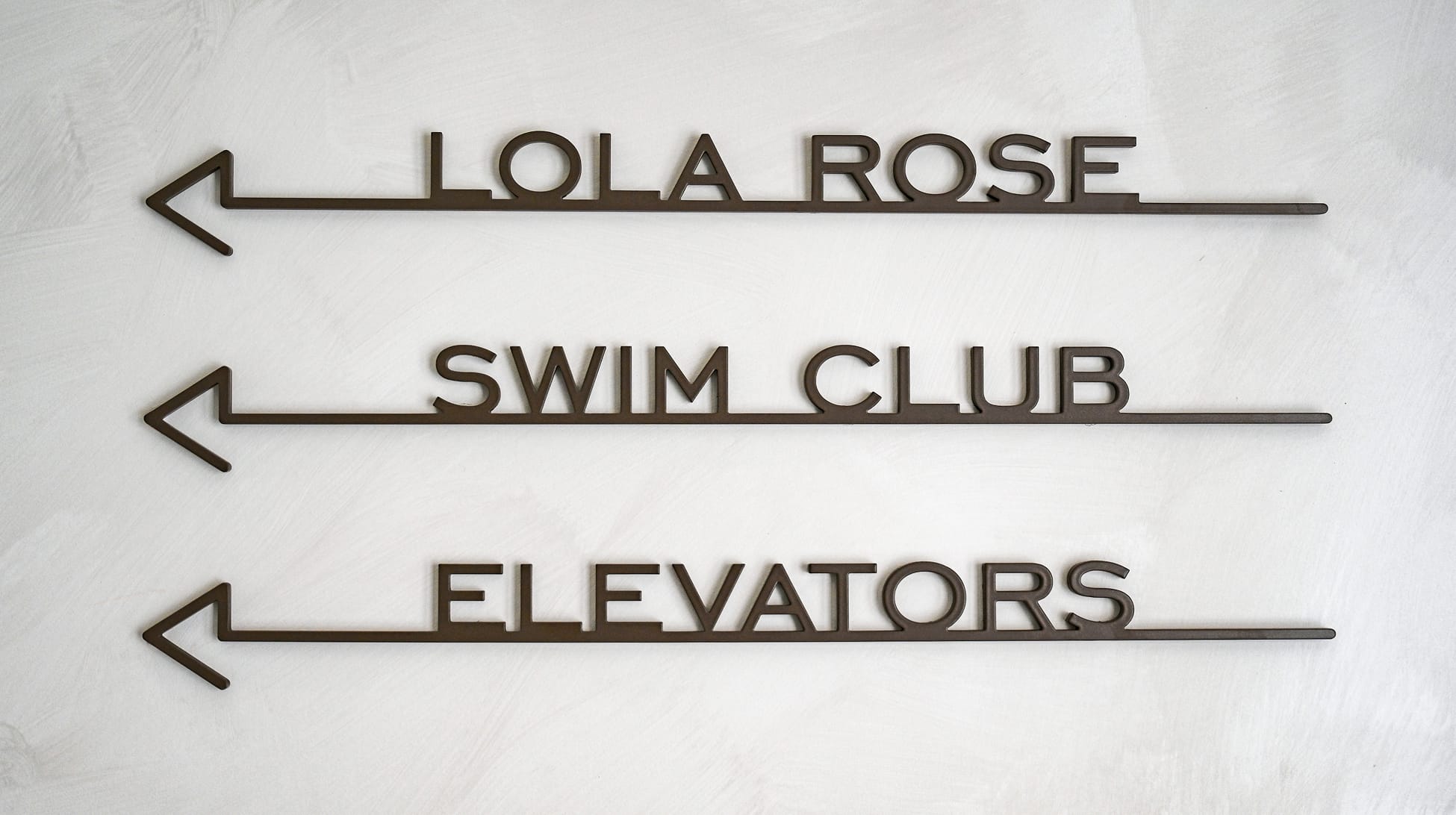 Three dark bronze metal directional wayfinding signs mounted on a white plaster wall at Thompson Palm Springs, with left-pointing arrows directing guests to Lola Rose, Swim Club, and Elevators.