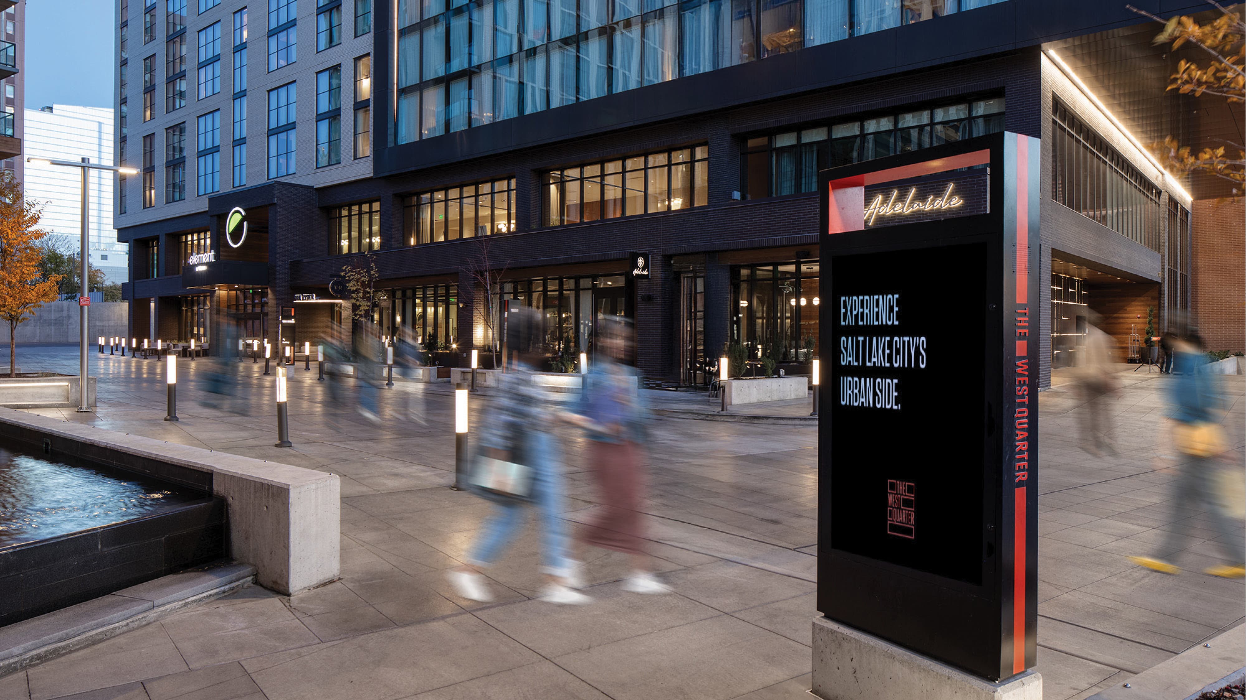 RSM Design digital integration and placemaking for The West Quarter in Salt Lake City, featuring a branded black and red digital display totem for The West Quarter and Adelaide restaurant positioned at the edge of an illuminated pedestrian plaza at dusk, with Element Hotel building signage, a water feature, bollard lighting, and motion-blurred pedestrians activating the public realm, illustrating how environmental graphic design, digital integration and interactive solutions, wayfinding and signage design, and branded environments work together to unify identity across a mixed-use hospitality, food and beverage, residential community, and urban streetscape development.