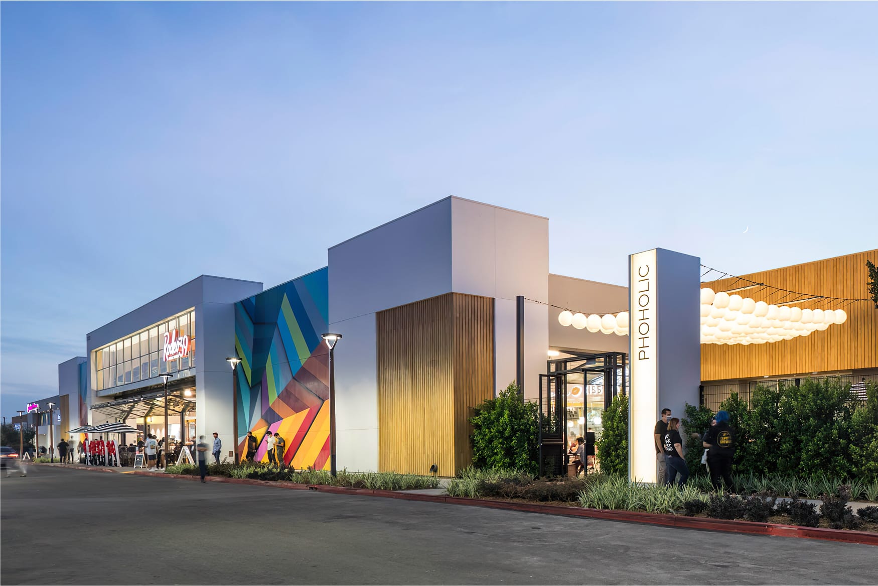 Exterior view of Rodeo 39 Public Market at dusk in Stanton, CA, featuring a vibrant geometric mural in bold colors, modern white and wood-clad facades, string lights, globe pendant lighting, and a illuminated vertical "Phoholic" sign, with guests gathered outside along the streetfront.