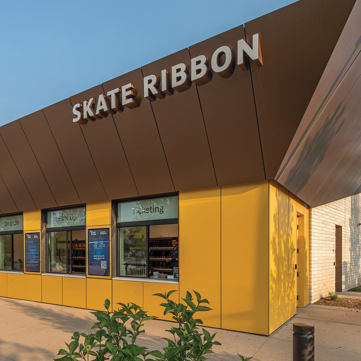 Skate Ribbon ticketing and skate rental building at Gene Leahy Mall RiverFront in downtown Omaha, Nebraska, featuring bold dimensional signage, a vibrant yellow facade, drop-off, pick-up, and ticketing windows against a clear blue sky