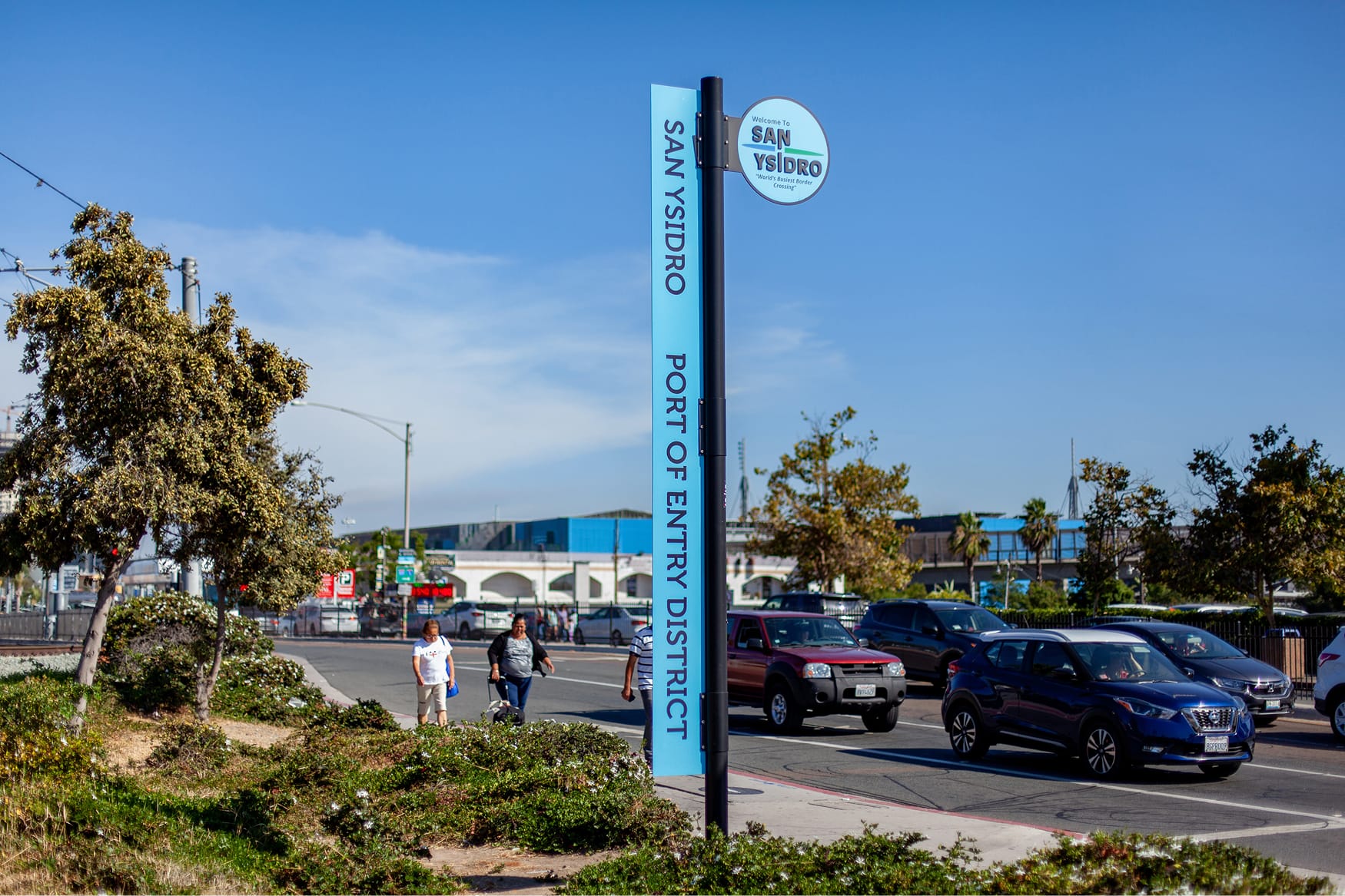 Blue vertical street sign reading 'San Ysidro Port of Entry District' with a circular emblem welcoming visitors to San Ysidro as the 'World's Busiest Border Crossing,' positioned beside a busy roadway with pedestrians and traffic.