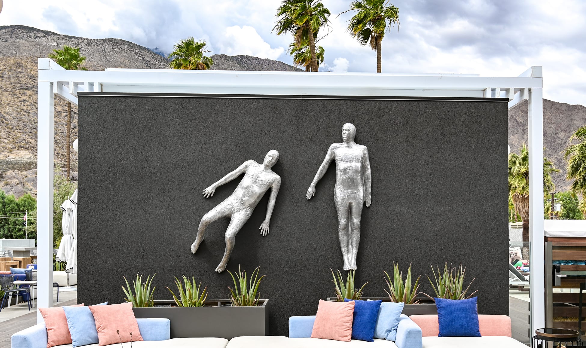 Two silver figural relief sculptures mounted on a dark accent wall at Thompson Palm Springs, set within a white pergola structure, with outdoor lounge seating, aloe planters, palm trees, and the San Jacinto Mountains in the background.