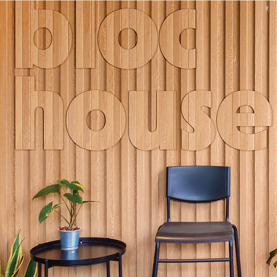 Interior photo of a Bloc House common area, showing the oversized "bloc house" wordmark routed directly into warm oak slatted wall paneling. A black metal side table with a potted plant and a black chair are positioned in front of the feature wall.
