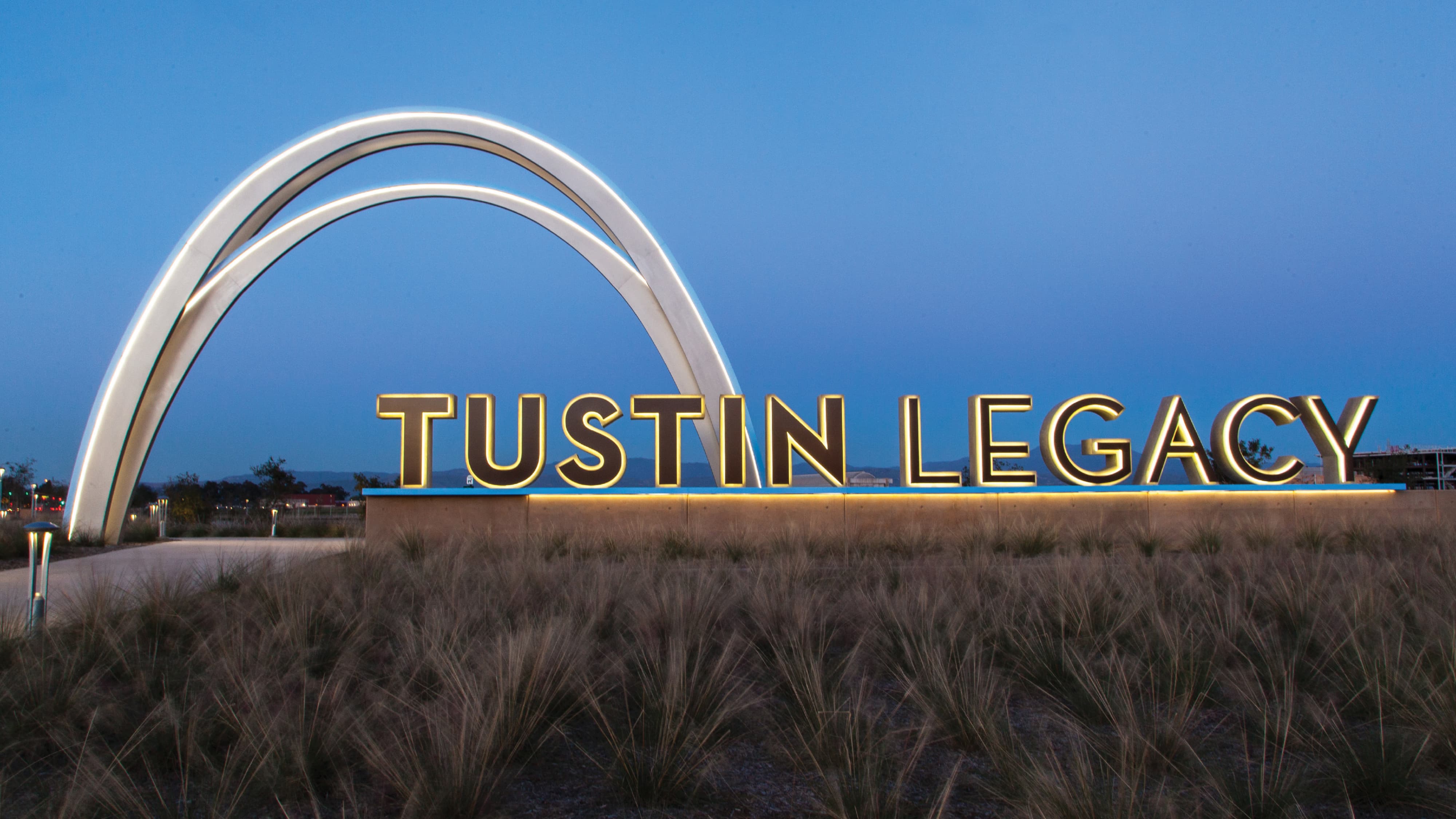 Illuminated 'Tustin Legacy' sign with backlit gold letters at dusk, set beneath a glowing white arched sculpture, surrounded by native grasses under a deep blue evening sky.