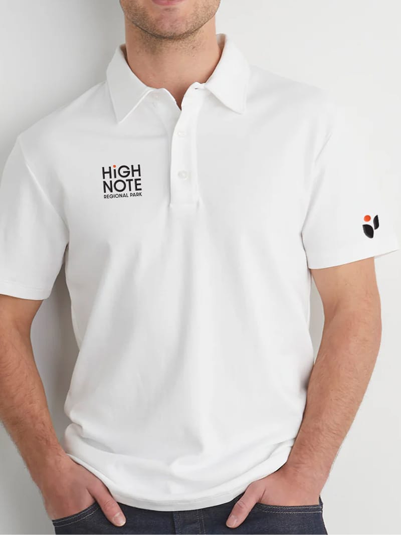 White polo shirt mockup on a model, embroidered with the "HighNote Regional Park" logo on the chest and the leaf-and-dot icon on the sleeve — brand identity and merchandise design by RSM Design.