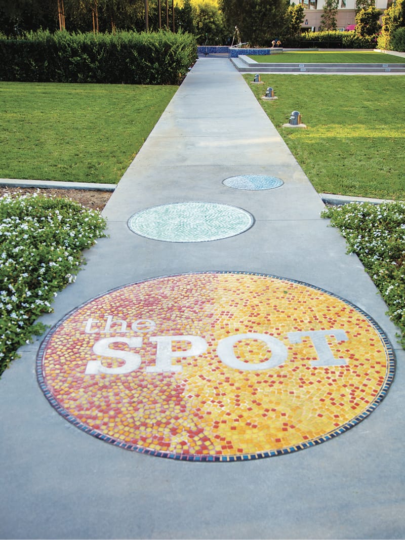 Circular mosaic tile medallion reading "the SPOT" in warm red and orange tones, designed by RSM Design, embedded in a concrete pathway leading through a lawn area in Playa Vista.